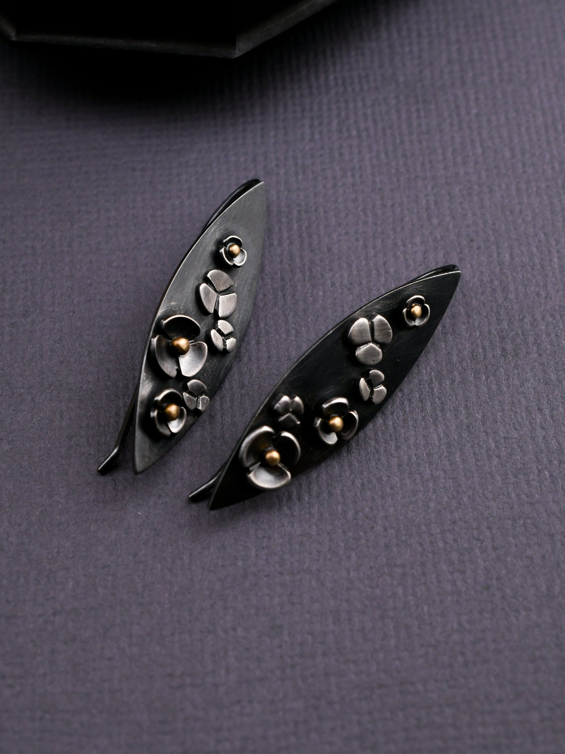 Leaf shaped earrings of blackened silver with floral details and gold accents on a dark textured background