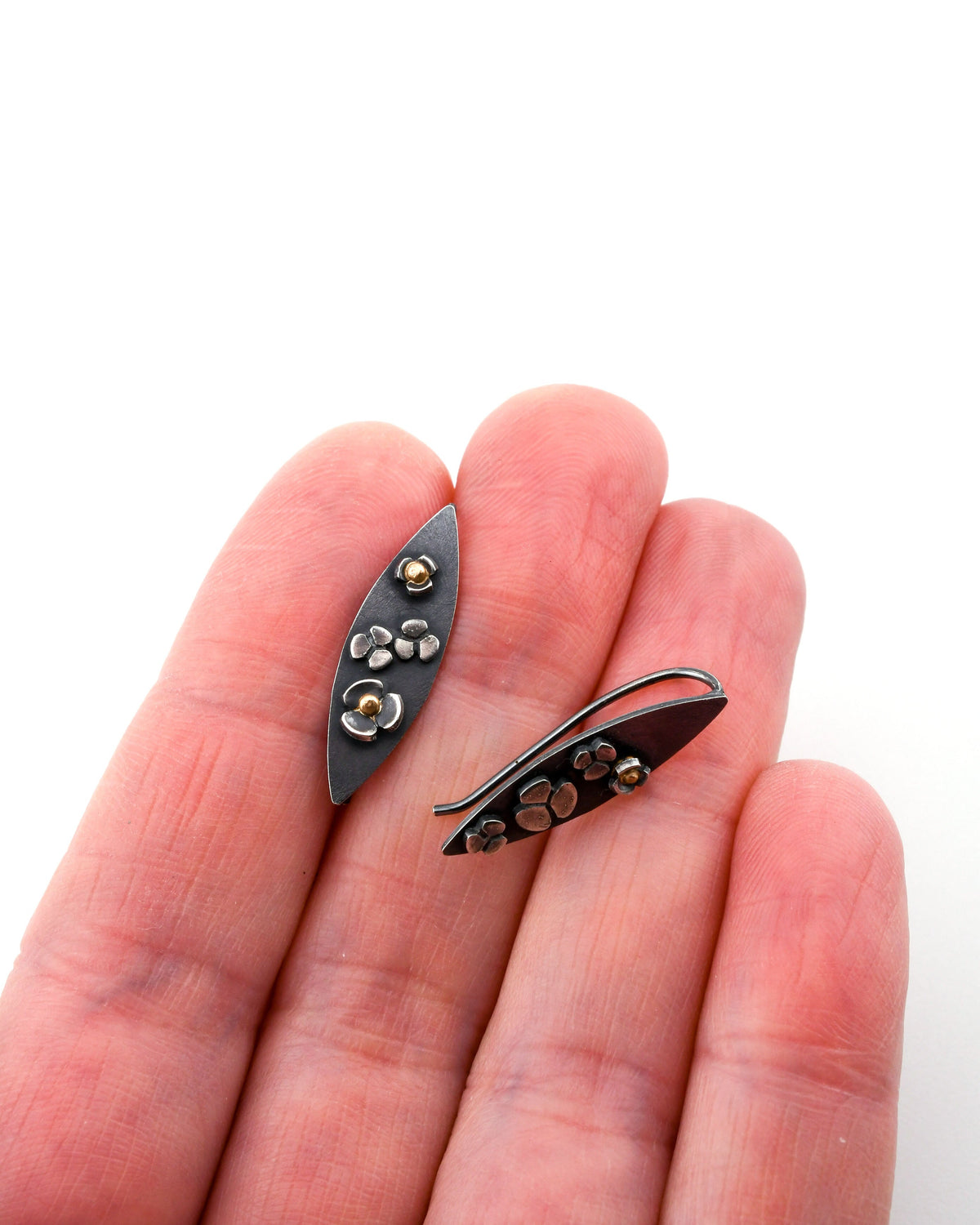 A pair of leaf-shaped blackened silver earrings with drifting flower designs held on a hand over a white background