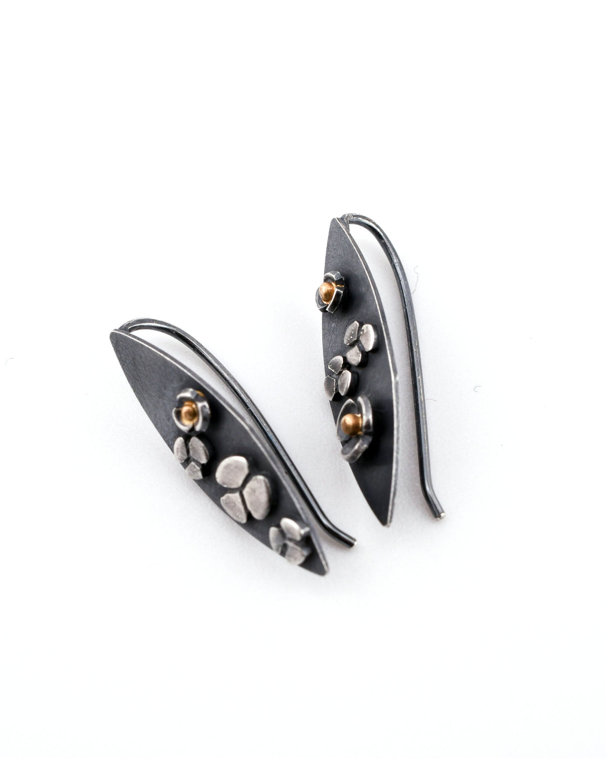 A pair of leaf-shaped blackened silver earrings with flower designs and 14k gold accents shown on their sides on a white surface