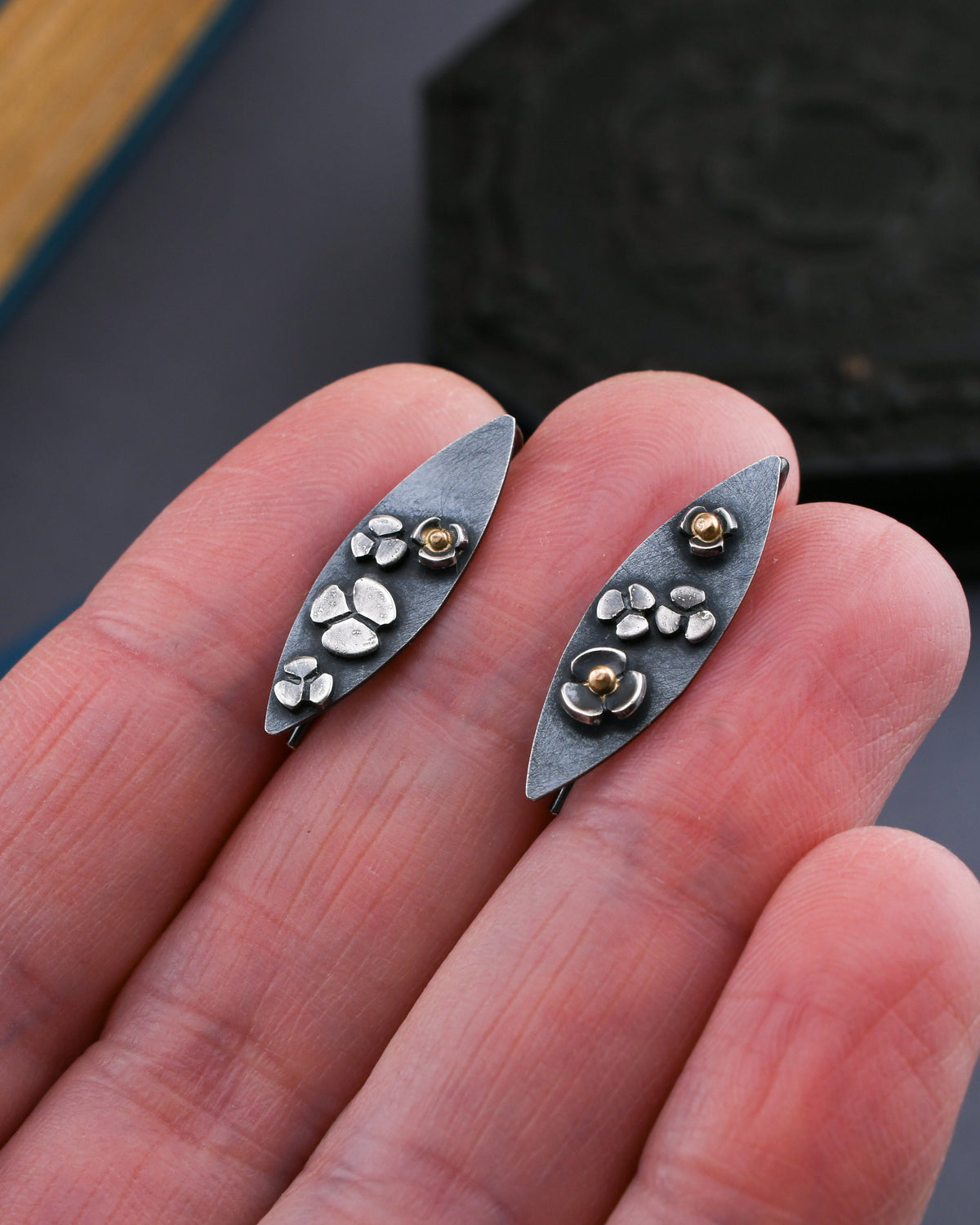 Silver earrings with floral designs and 14k gold accents held between fingers against a blurred background