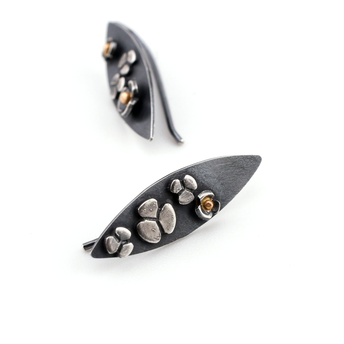 A pair of leaf-shaped earrings with stylized three-lobed flowers  showing 14k gold centers in some of the flowers. The earrings are shown on their sides on a white surface