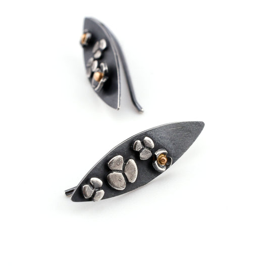 A pair of leaf-shaped earrings with stylized three-lobed flowers  showing 14k gold centers in some of the flowers. The earrings are shown on their sides on a white surface