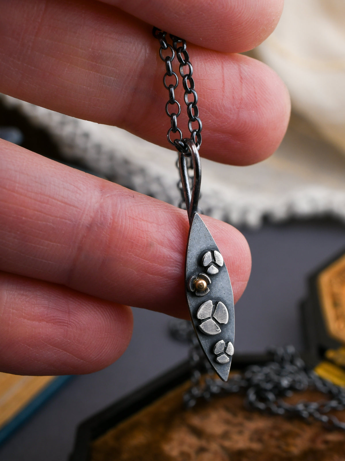 Hand holding a necklace with a leaf-shaped pendant featuring tiny flowers.