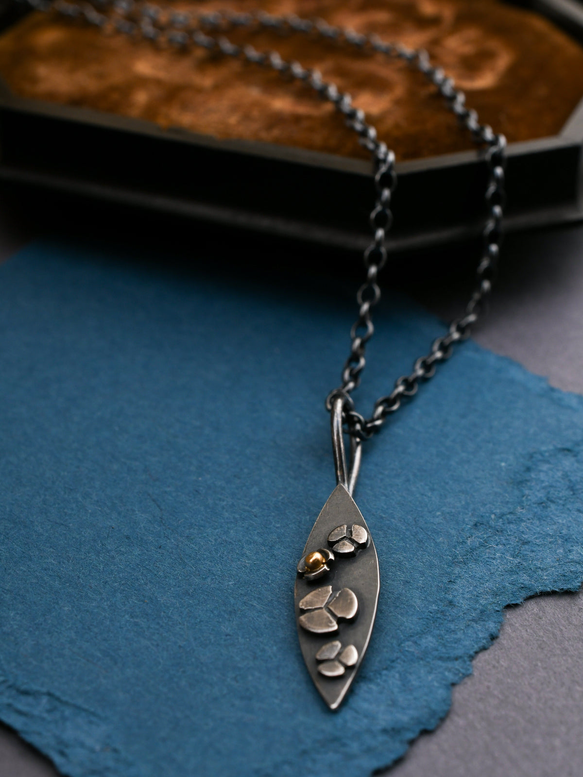 A blackened silver necklace with a leaf-shaped pendant showing stylized three-lobe flowers on the surface, displayed on a dark textured background with a brown velvet case behind