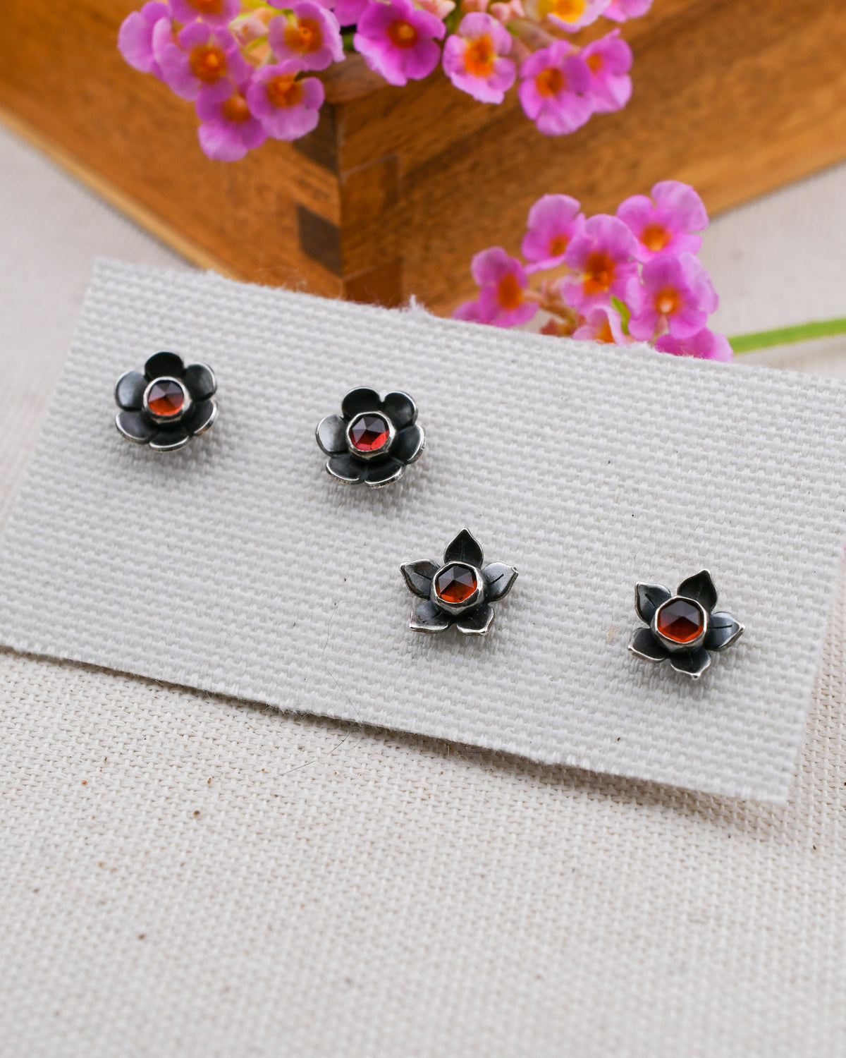 Two pairs of black flower-shaped stud earrings with red garnet centers, displayed on a white textured card with pink flowers and a wooden box in the background.