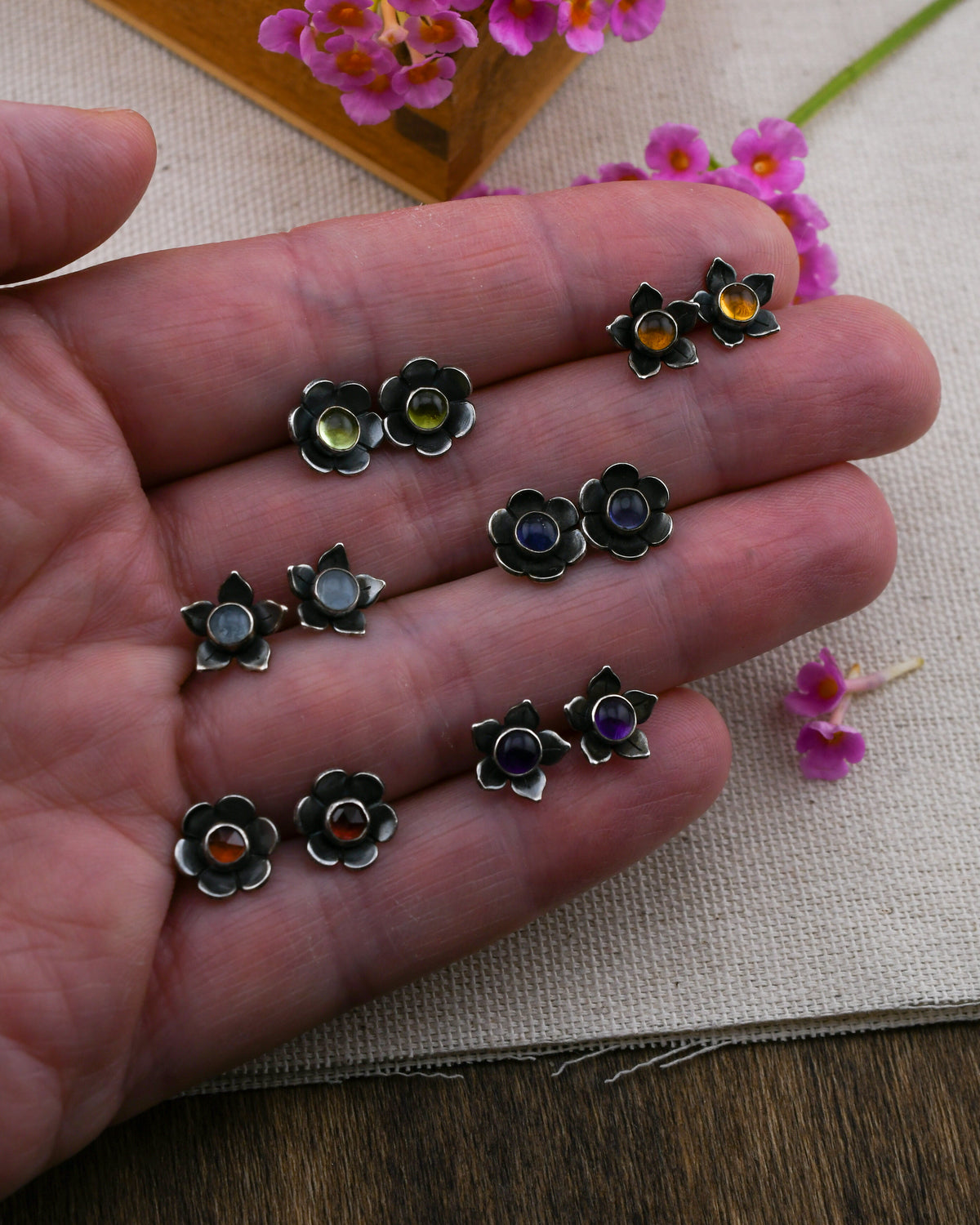 A hand holding several small flower-shaped stud earrings with dark metal petals and colorful round stones in yellow, green, blue, purple, and red, displayed over a textured fabric background with pink flowers.