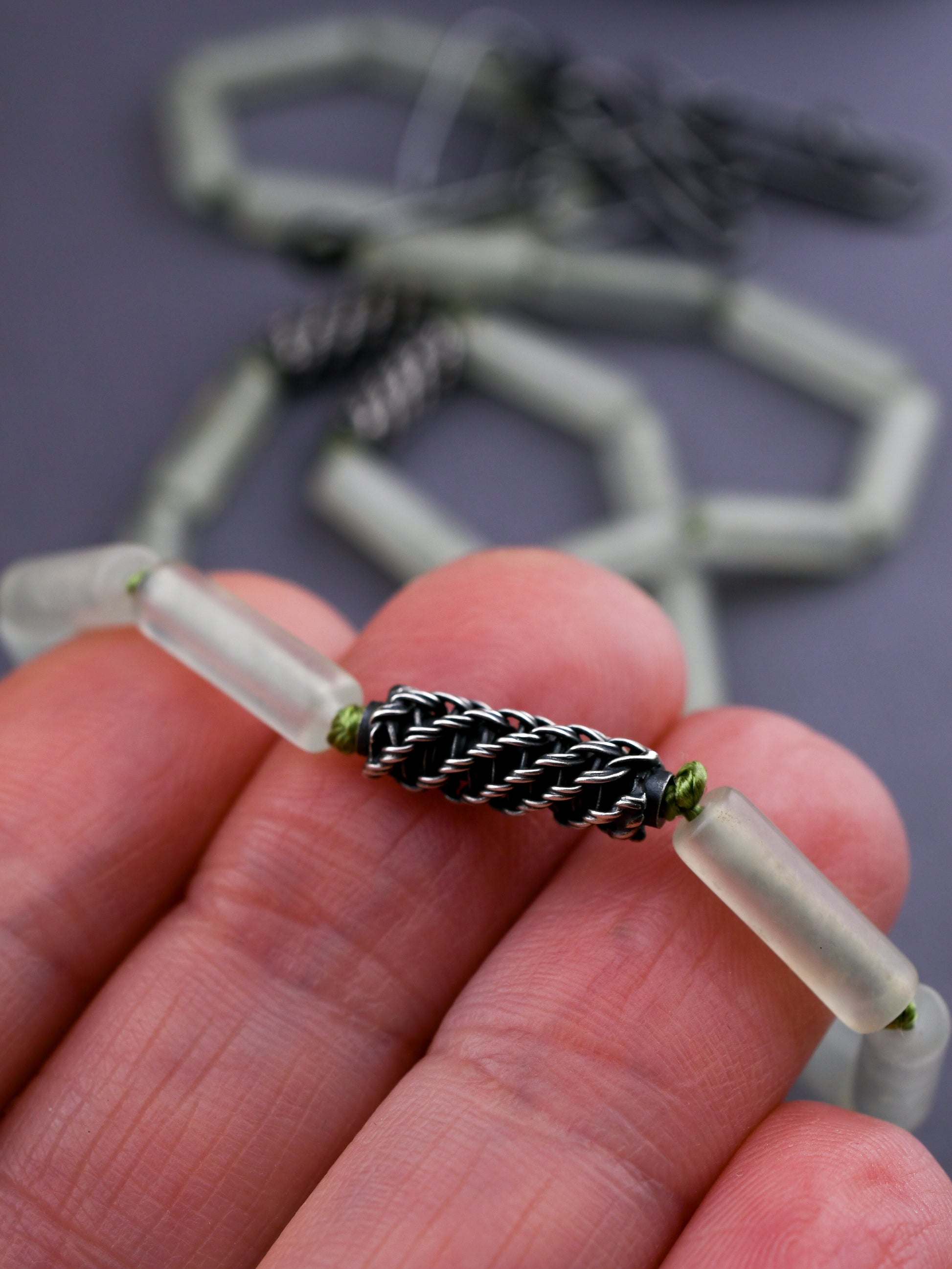 Close-up of a hand holding a woven silver bead and green stone bead over a dark blurred background