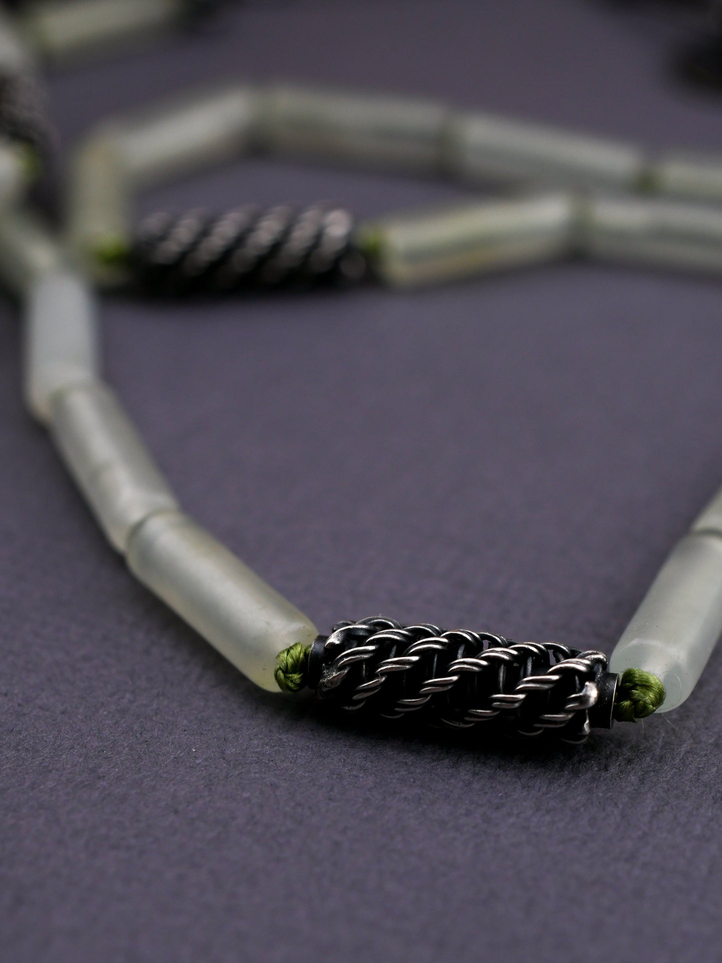 close-up of woven silver bead knotted with green silk cord and green stone beads on a textured gray background