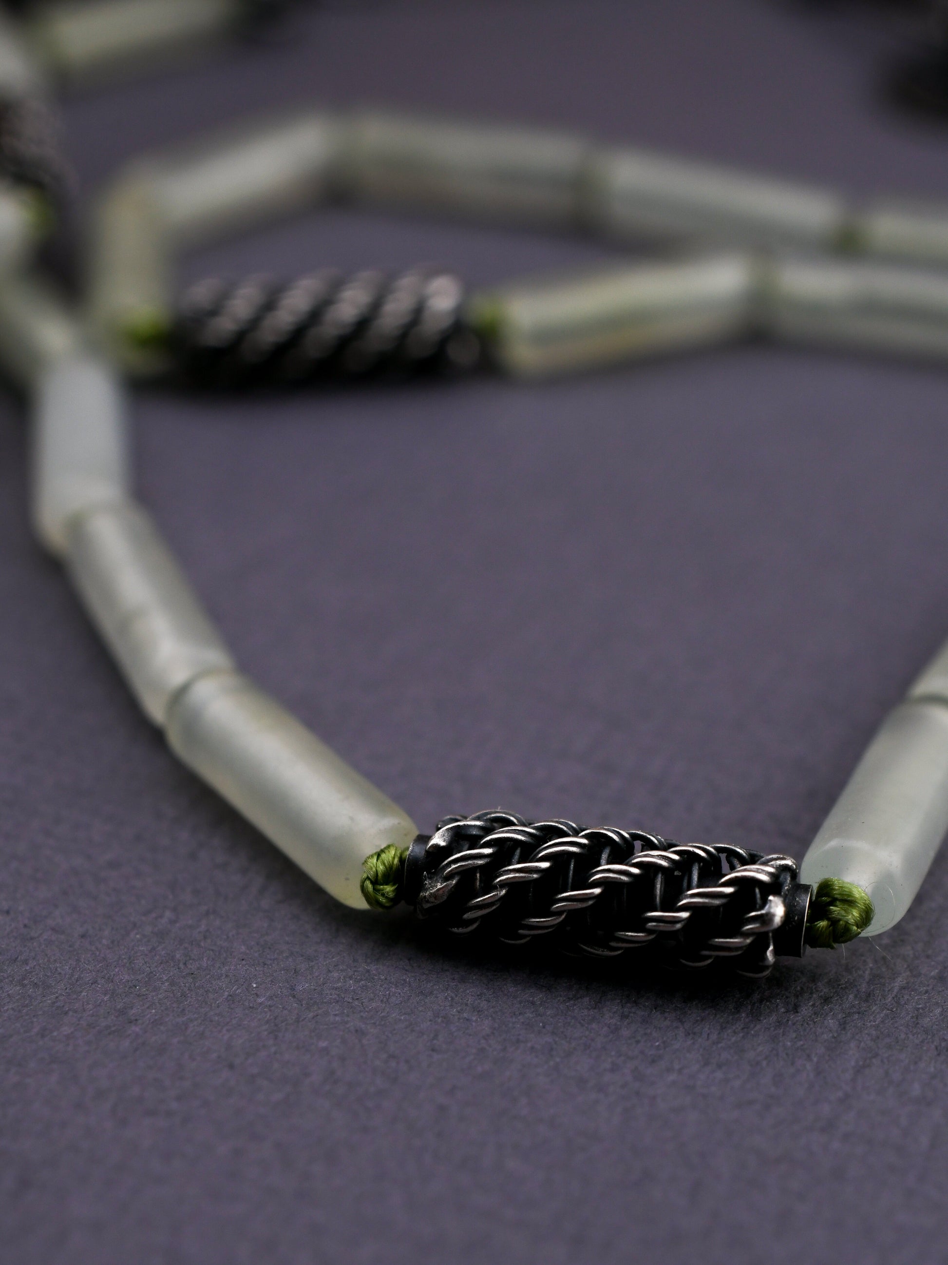 close-up of woven silver bead knotted with green silk cord and green stone beads on a textured gray background