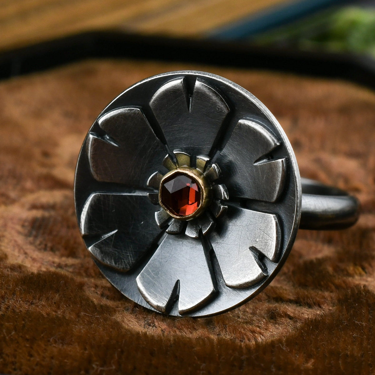 Helios ring in blackened sterling silver with garnet set in 14k gold displayed on brown velvet with a blurred background