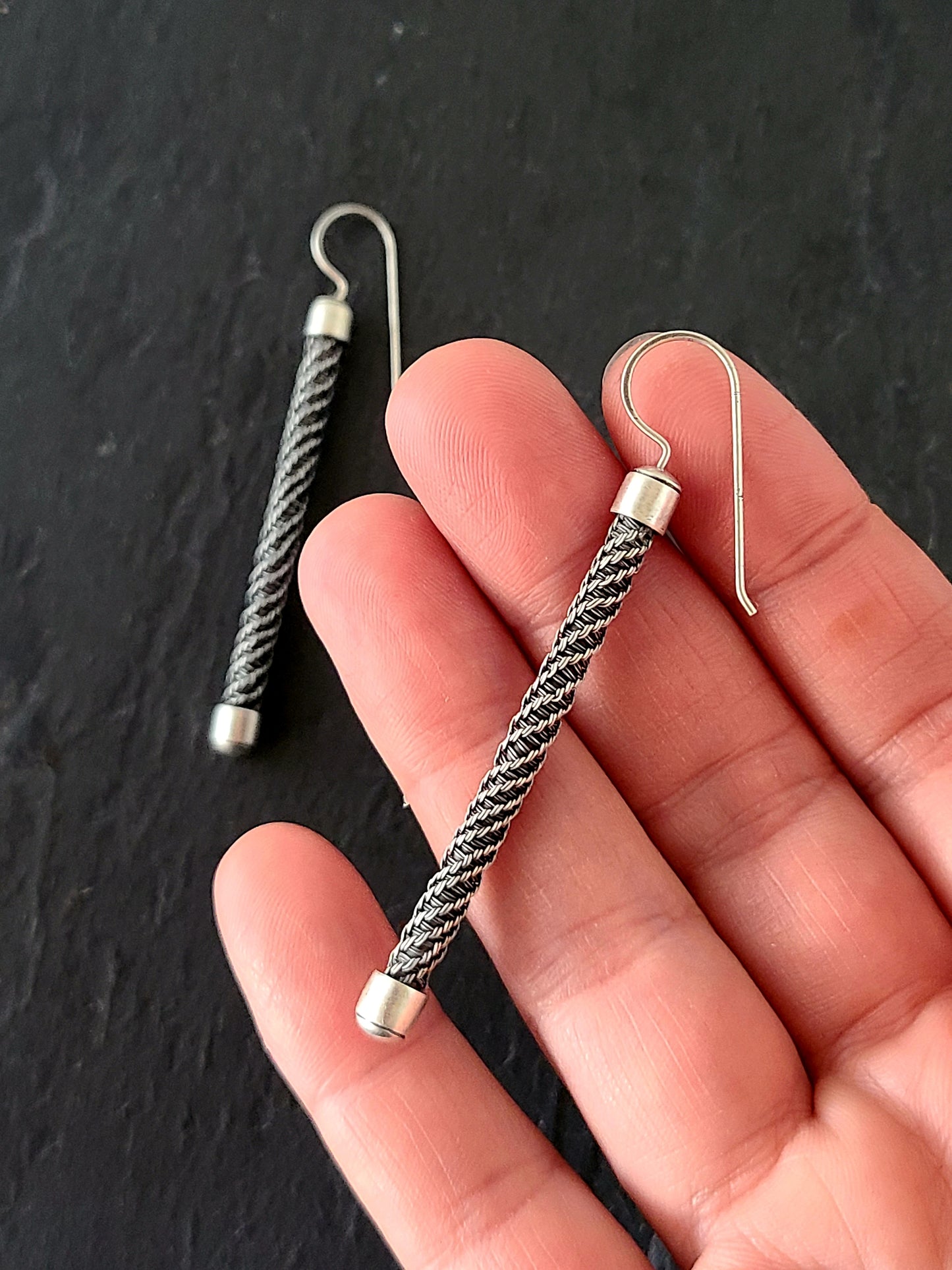 Long silver earrings with twisted wire pattern and bright silver end caps held in a hand on a black background