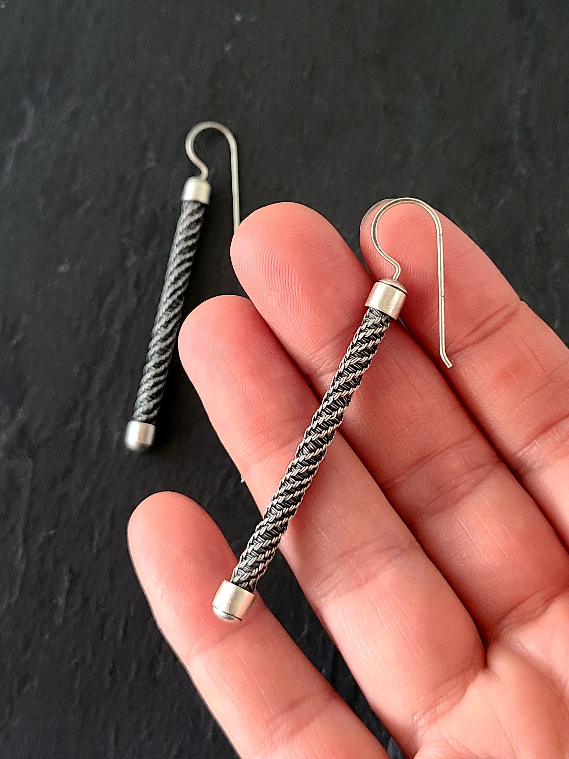 Long silver earrings with twisted wire pattern and bright silver end caps held in a hand on a black background