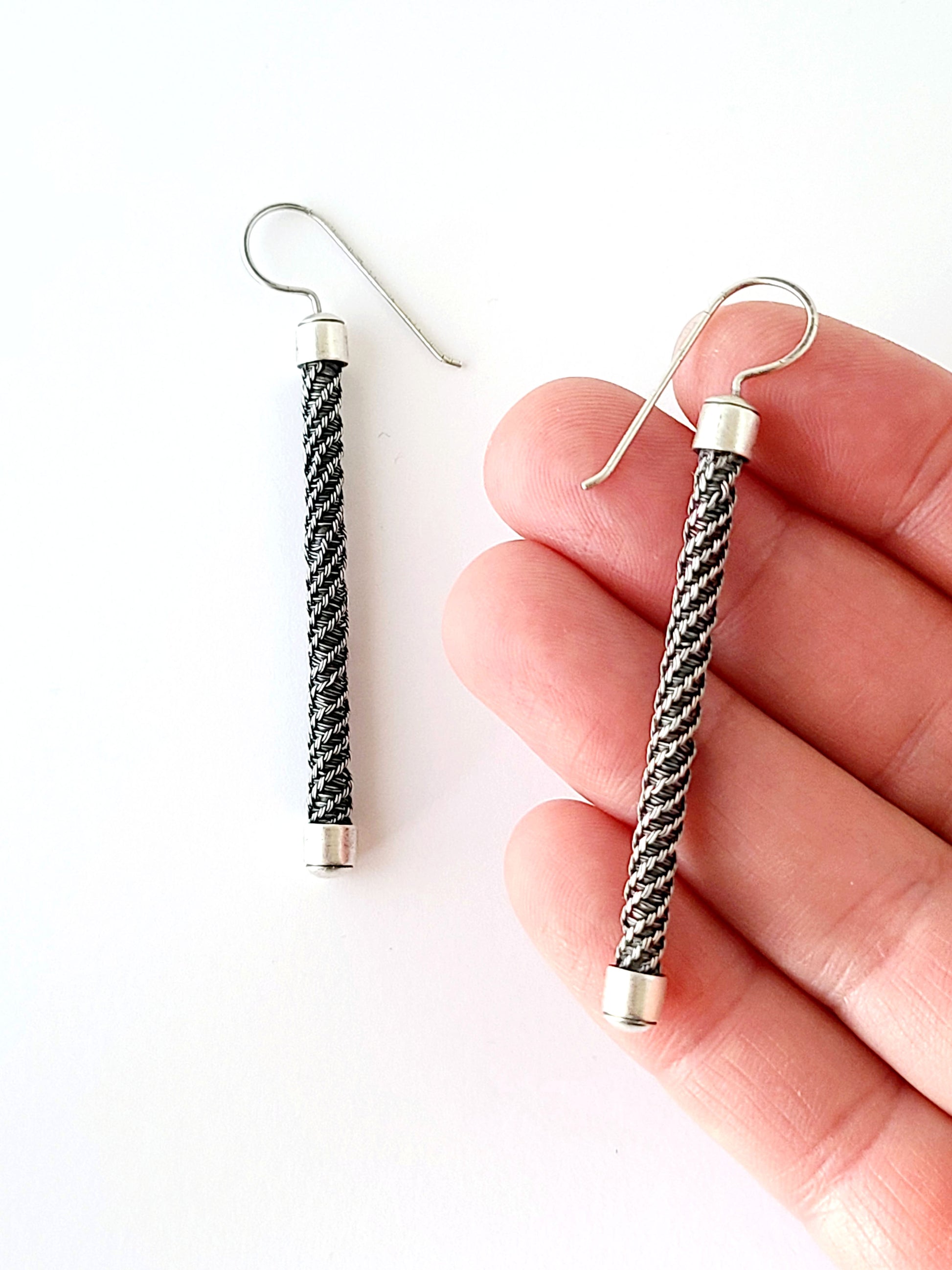 Pair of silver and black braided earrings held by a hand on a white background