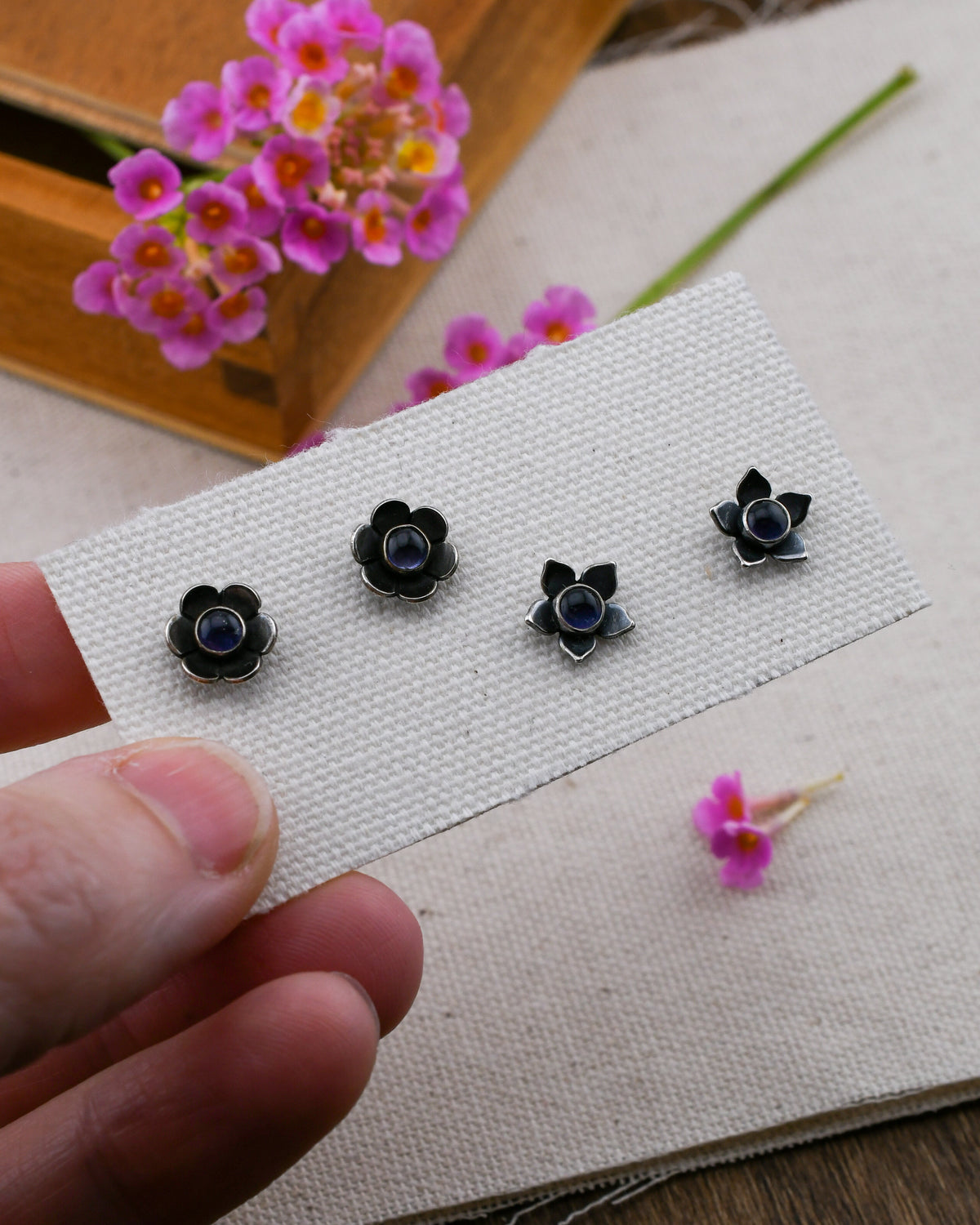 A hand holds a small white textured card displaying two pairs of black flower-shaped stud earrings with dark blue iolite centers, set against a soft beige fabric background with pink flowers and a wooden box.