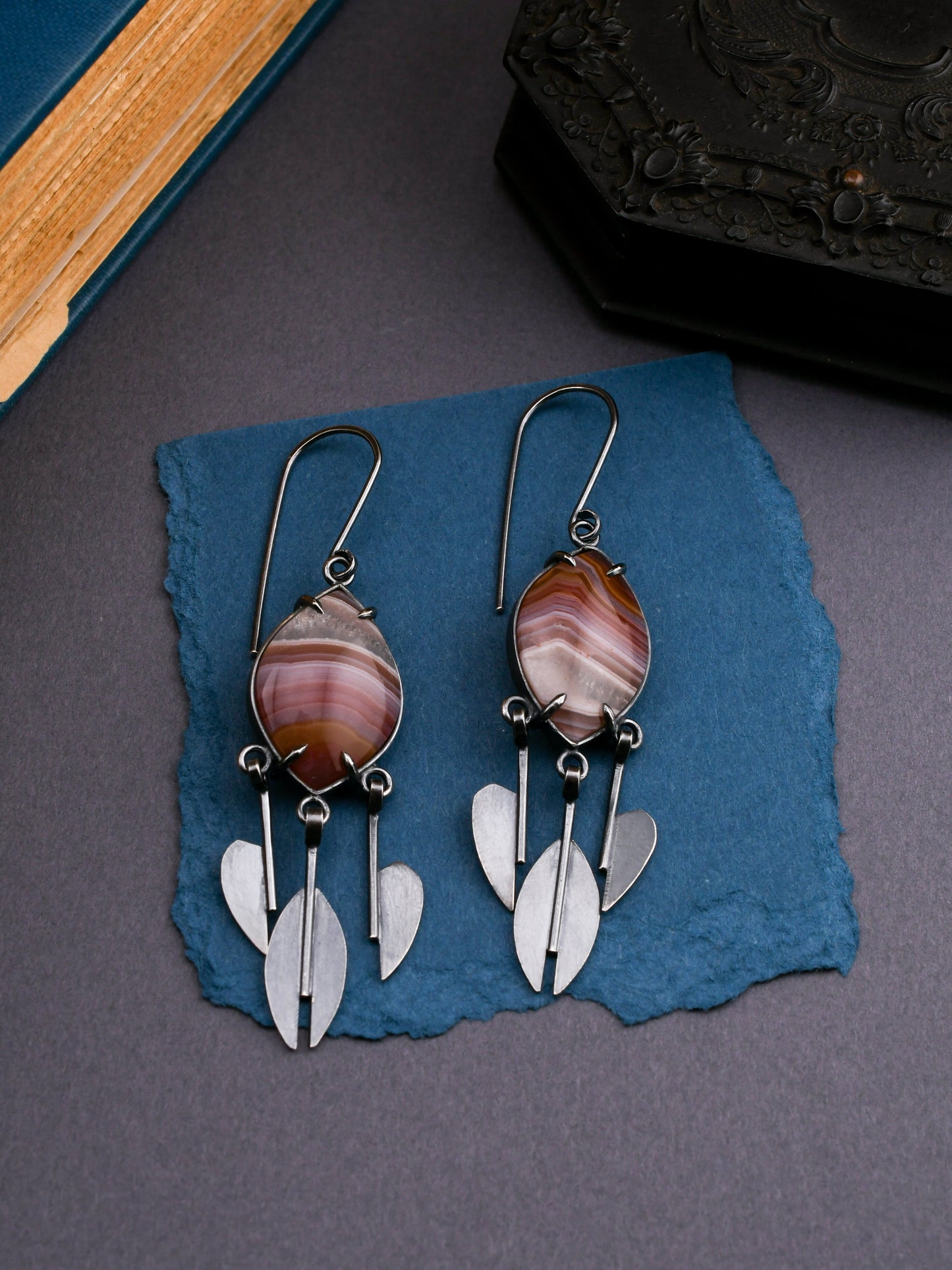 Pair of earrings with agate stones and silver raindrop pendants on a blue textured surface.