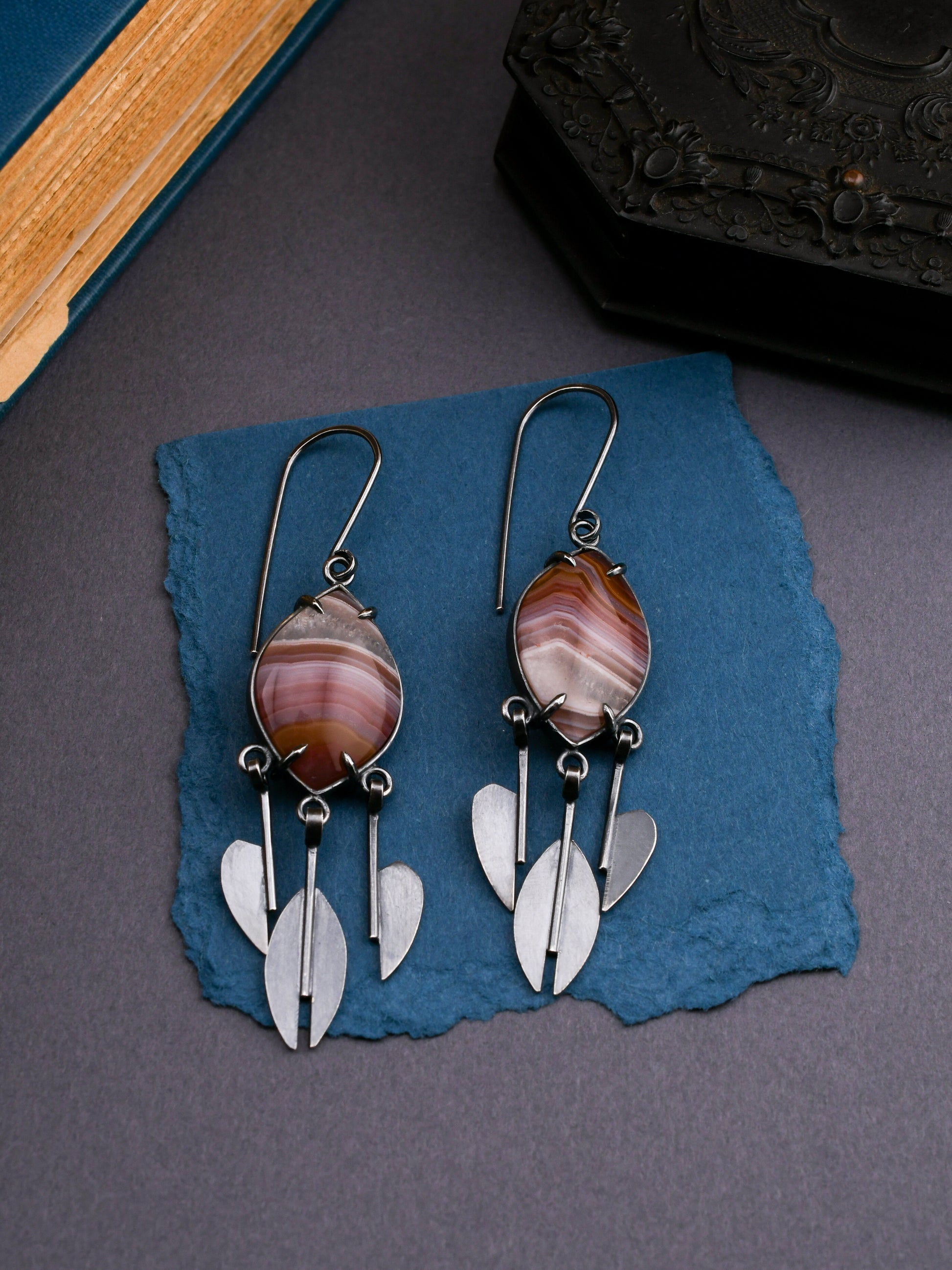 Pair of earrings with agate stones and silver raindrop pendants on a blue textured surface.