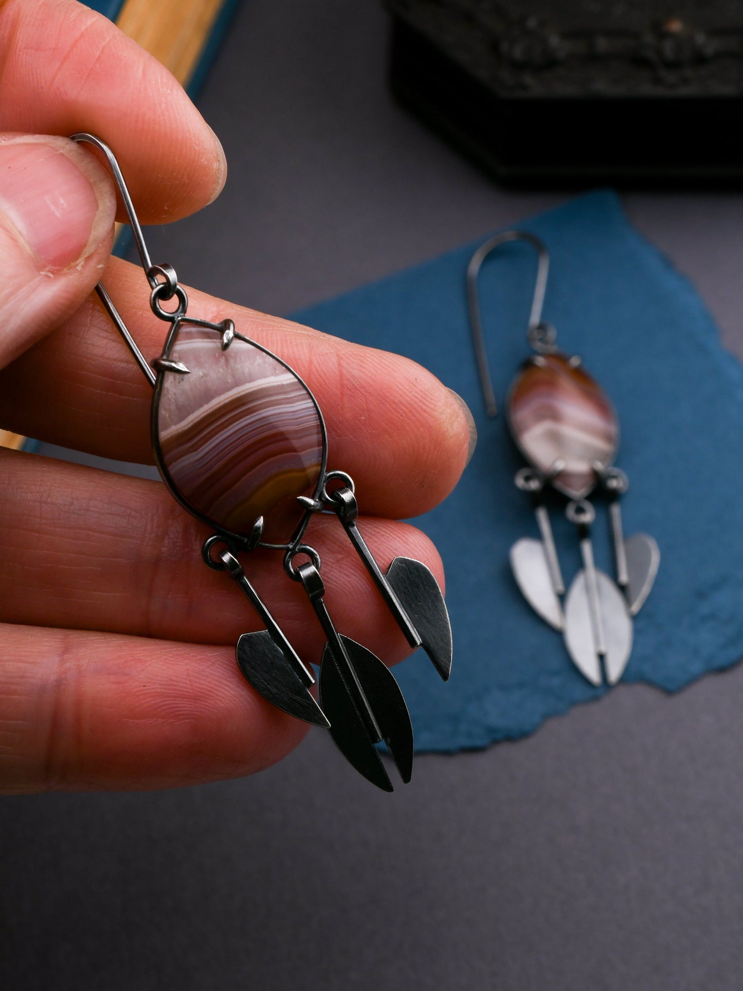Hand holding a pair of earrings with agate stone and raindrop design on a blue surface.