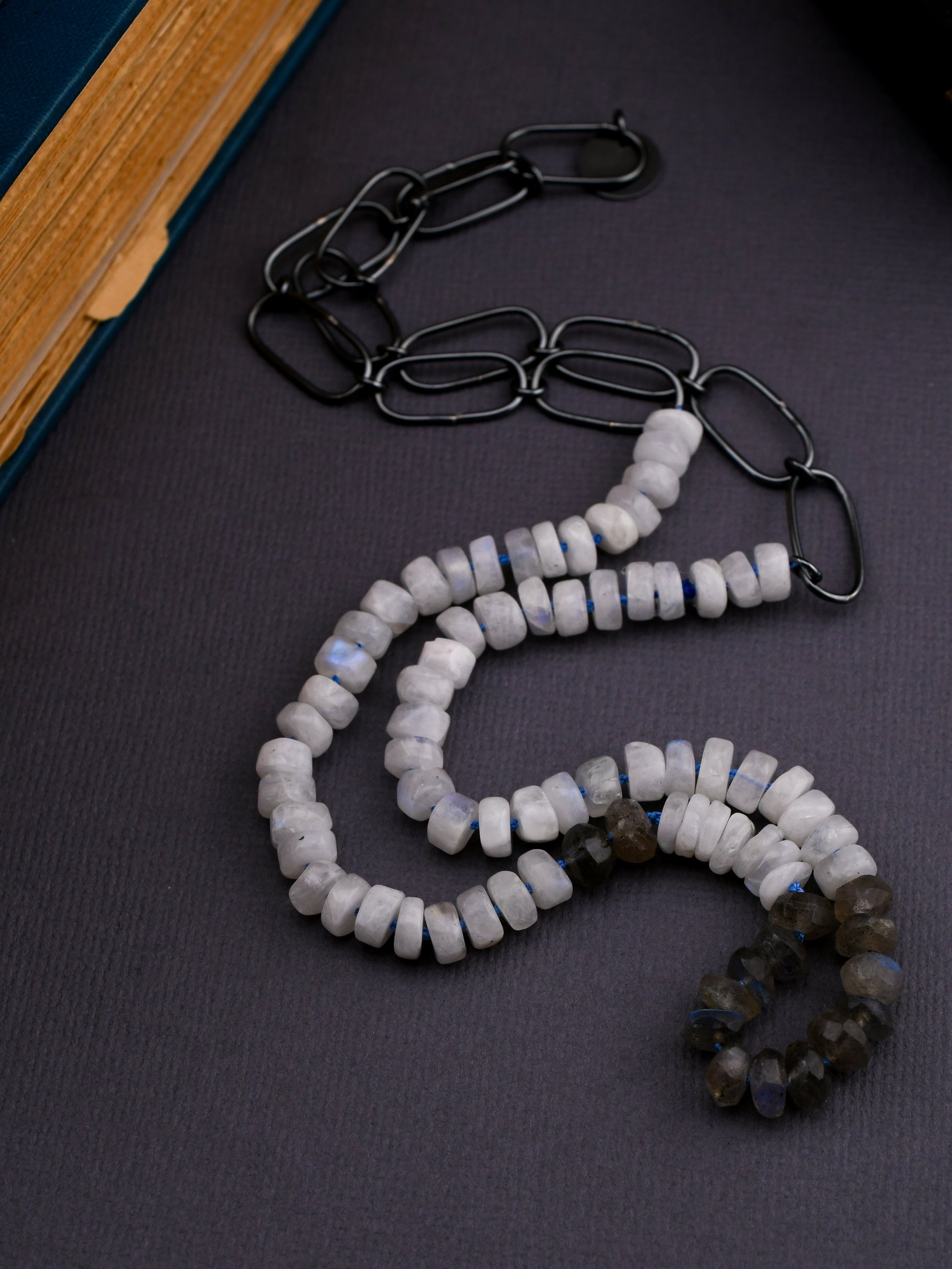 moonstone and labradorite bead necklace with blackened silver chain on a dark gray background next to a book