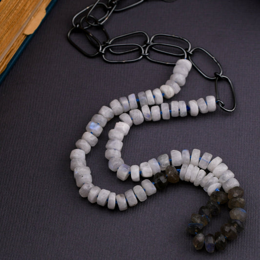 moonstone and labradorite bead necklace with blackened silver chain on a dark gray background next to a book