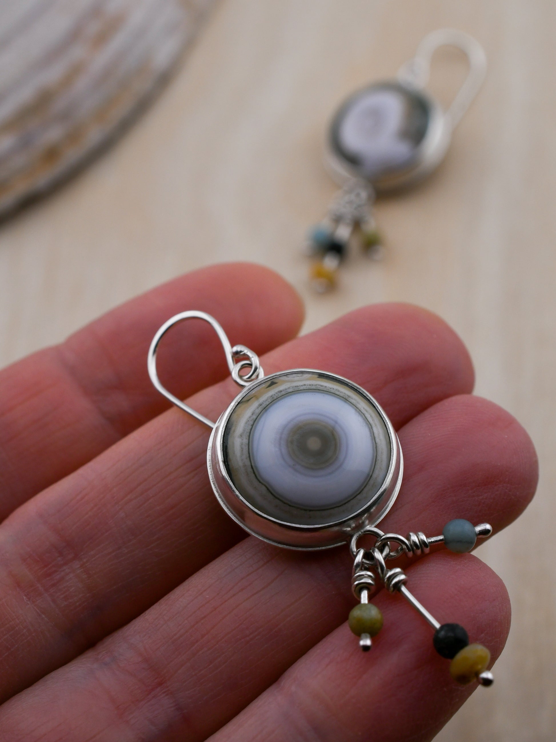 Close-up of a hand holding a silver earring with a round ocean jasper and beads.