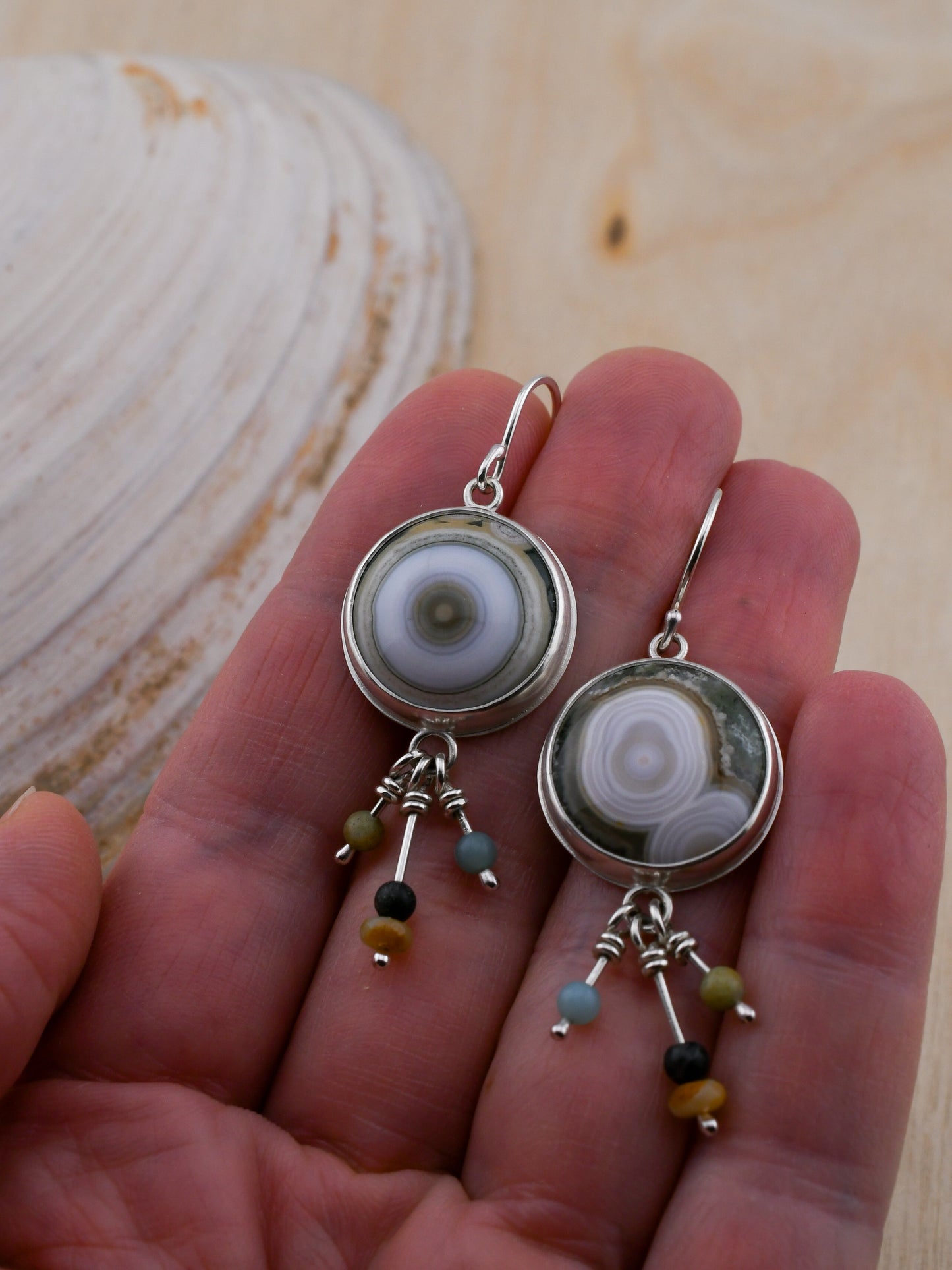 Silver earrings with round green and white ocean jaspers and colorful bead dangles held in a hand with a shell and wood background