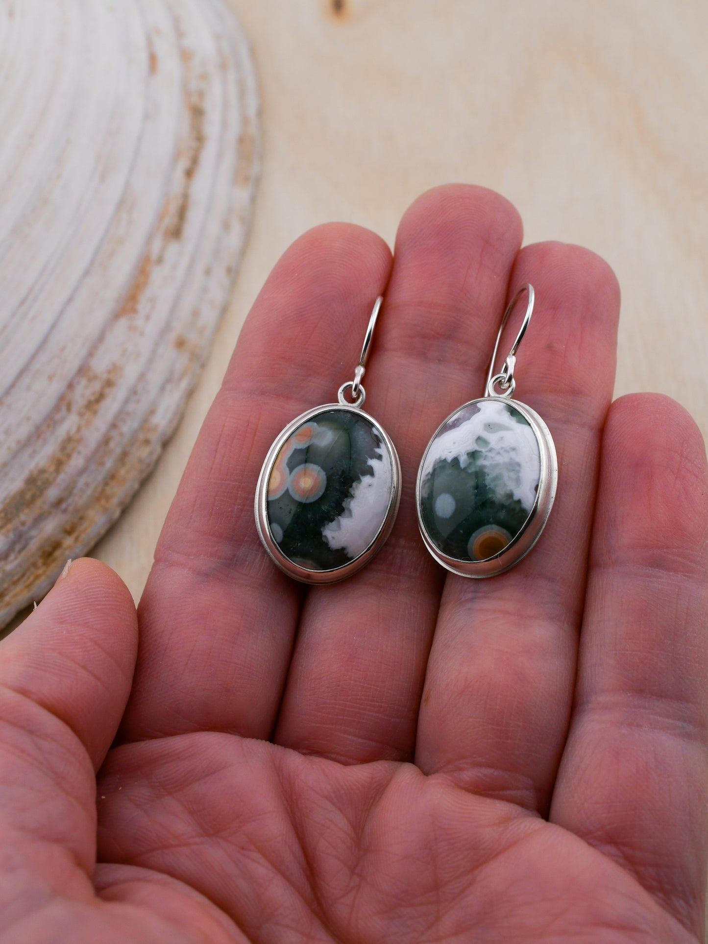 Silver earrings with green and white ocean jaspers held in a hand against a shell background