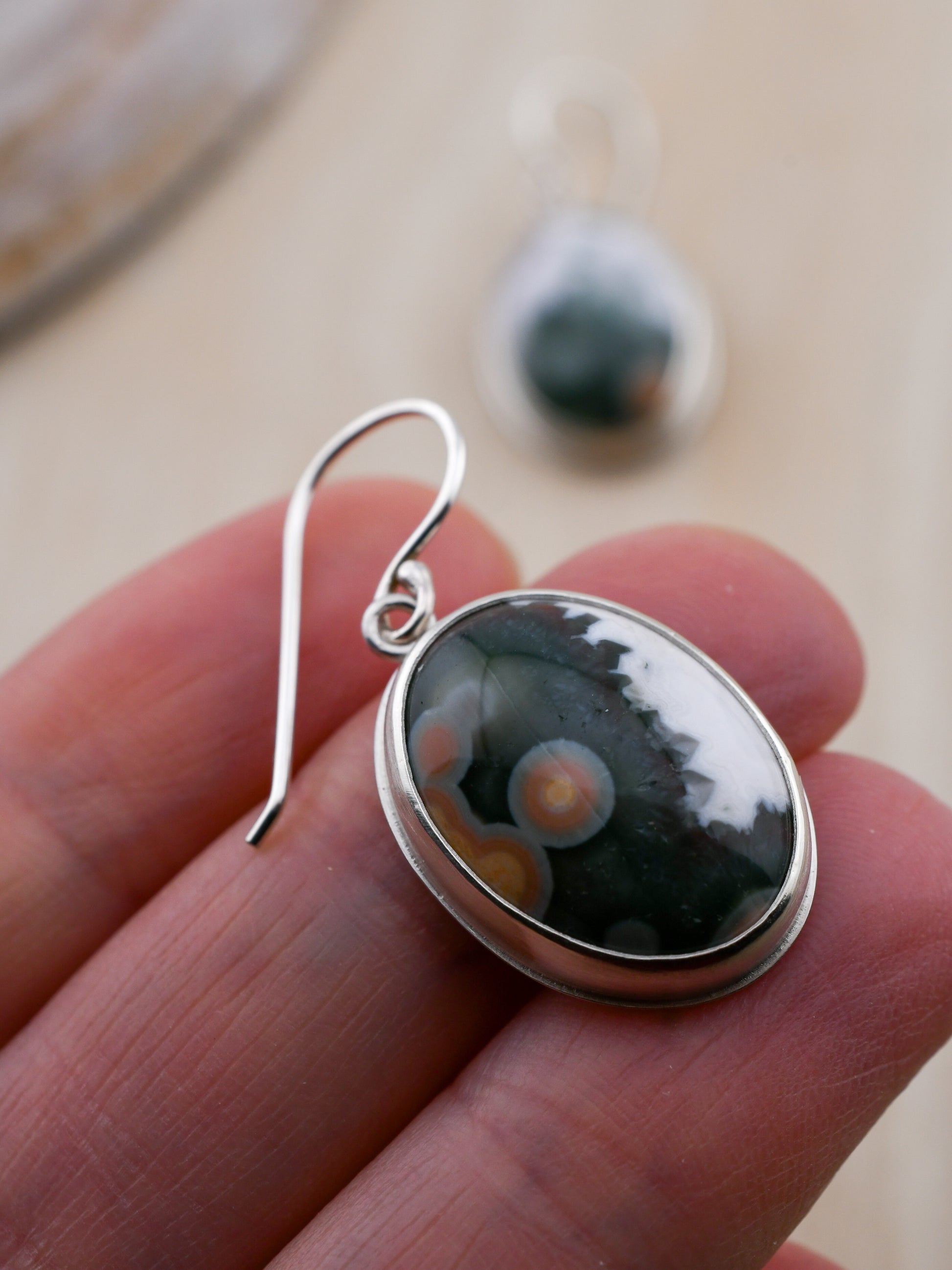 Silver earring with a green, white and orange speckled stone held between fingers on a blurred background