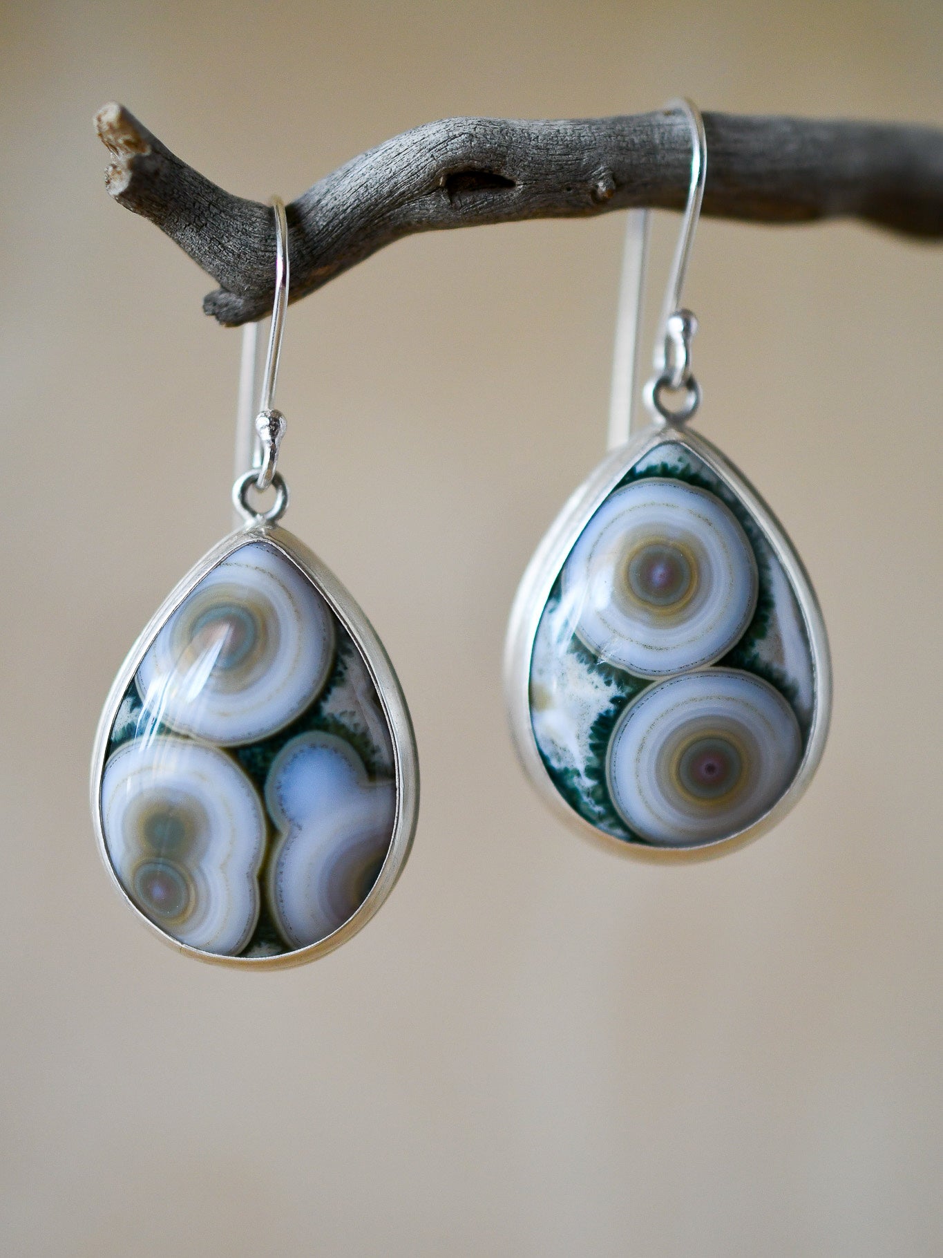 Teardrop-shaped earrings with intricate patterns on a branch against a beige background