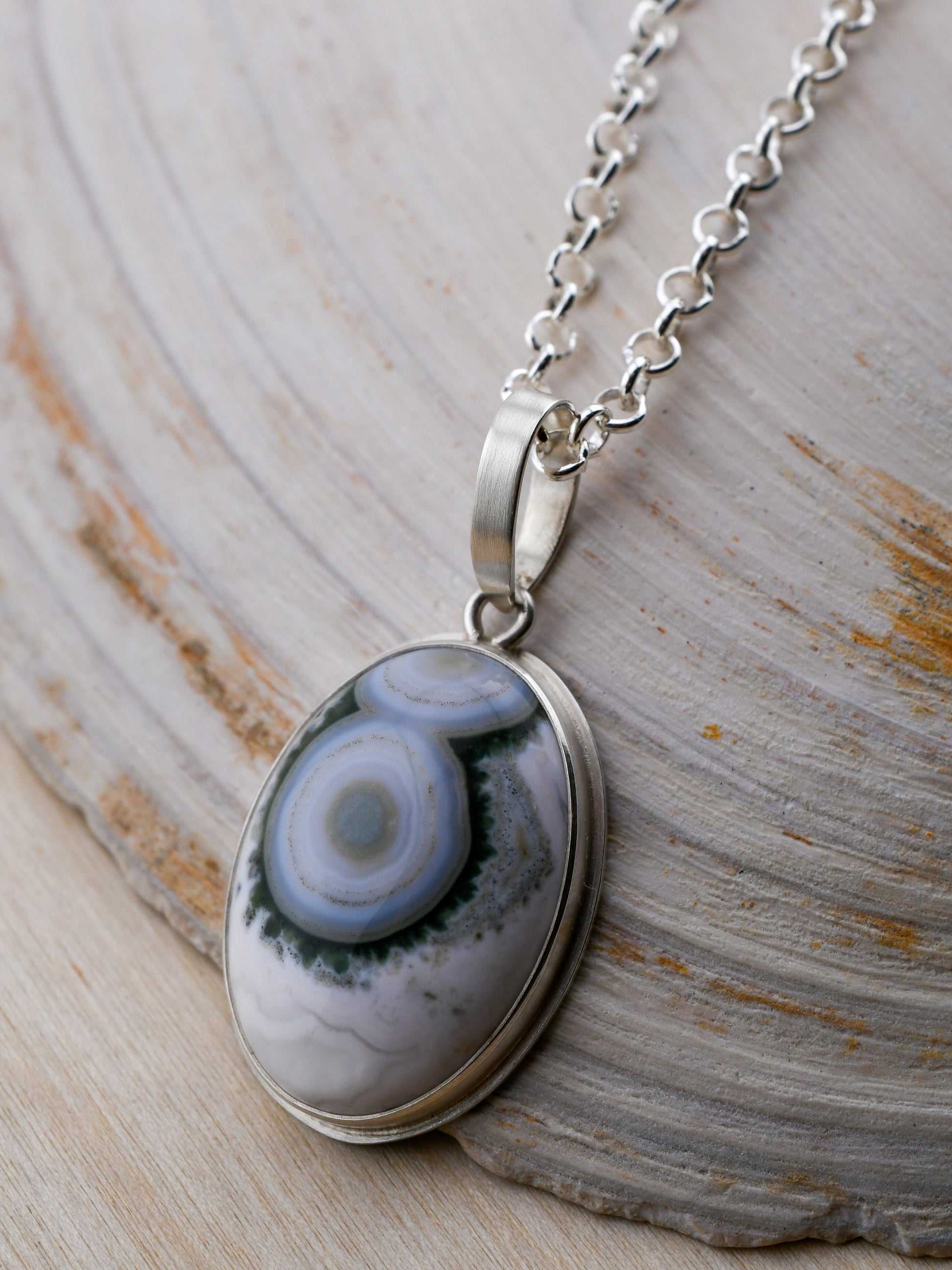 Silver necklace with green and white ocean jasper pendant displayed on a white shell and wood background