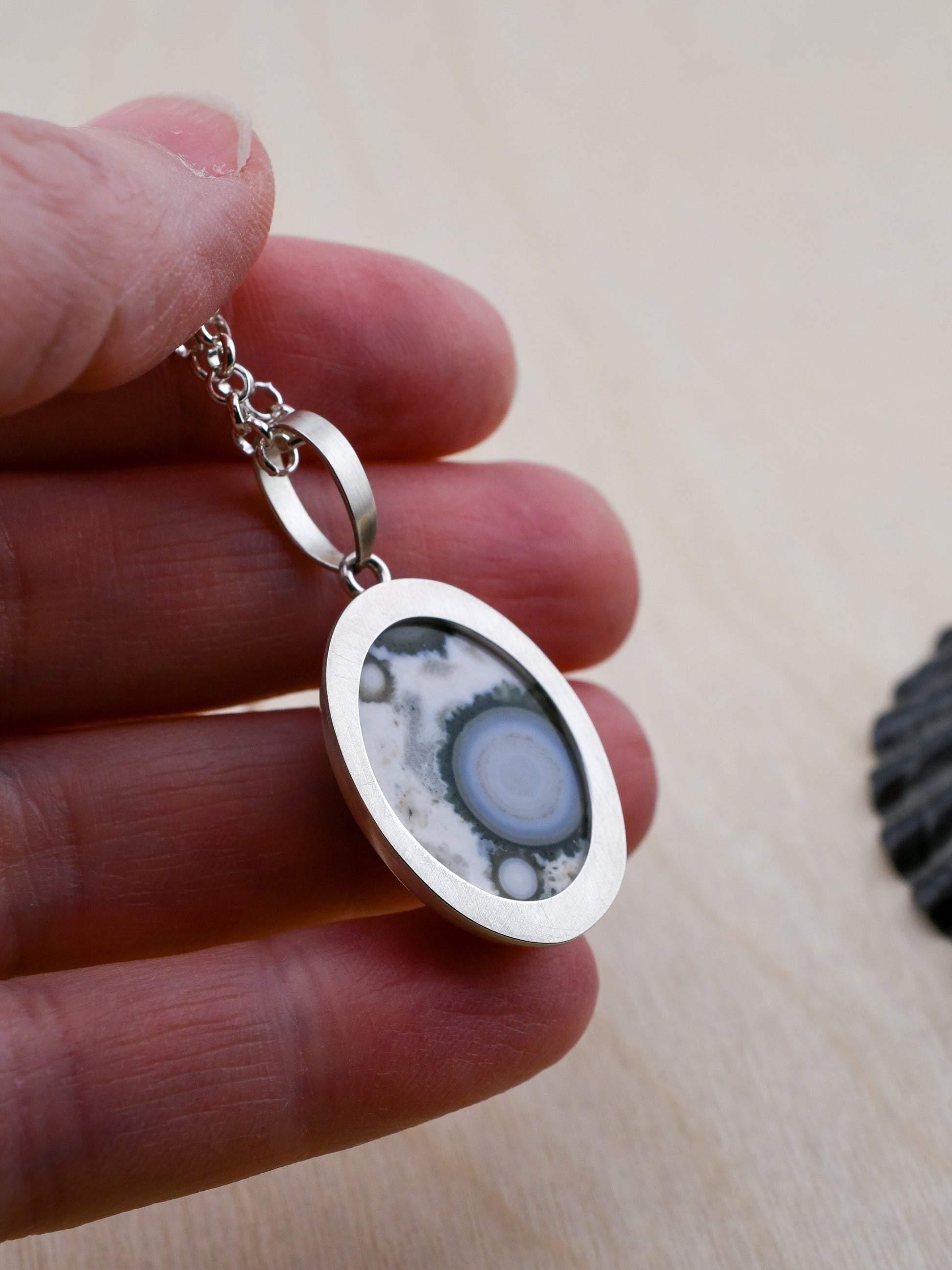 closeup of the backside of a silver pendant with a patterned stone held between fingers on a light wooden surface