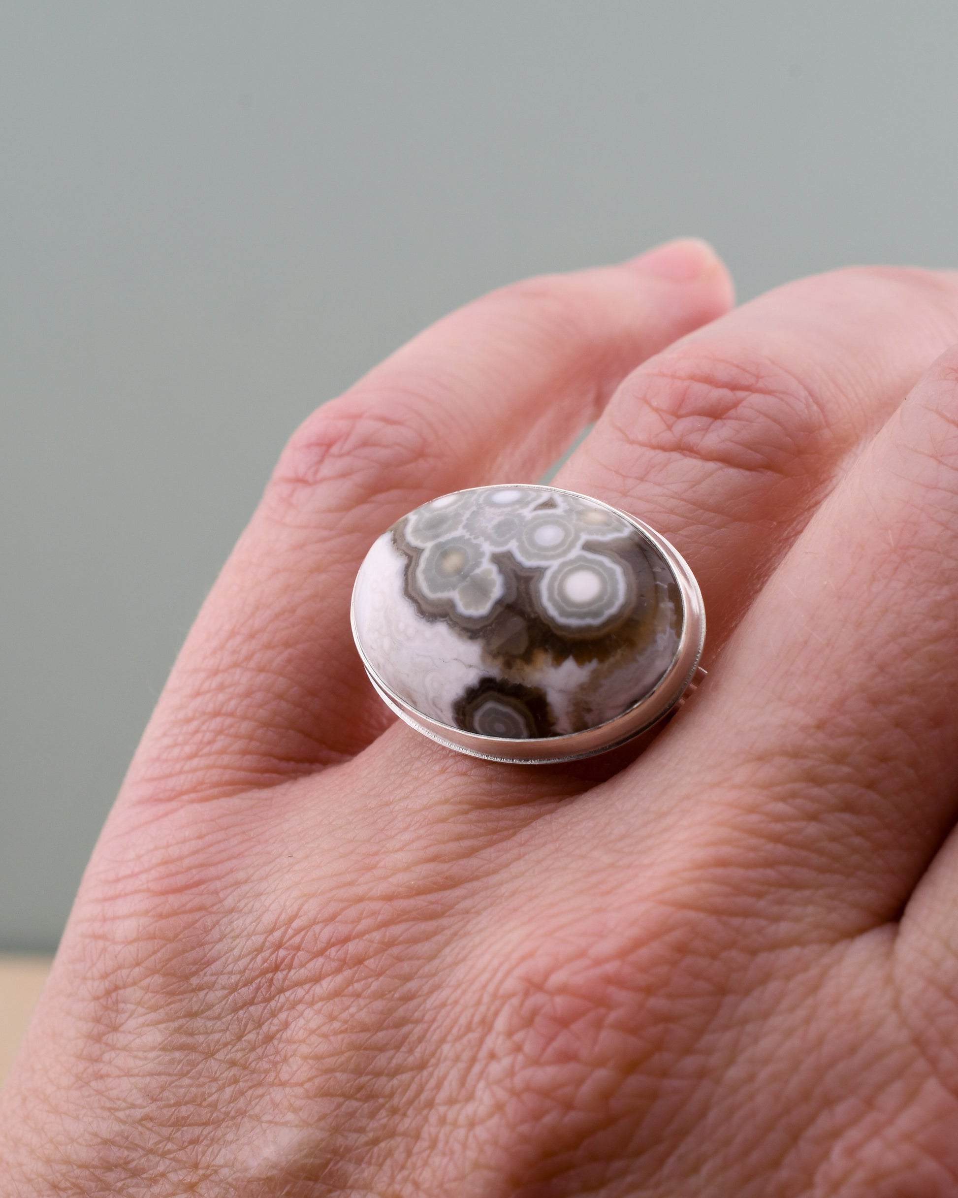 Hand wearing a silver ring with an oval brown and white ocean jasper with green circles on a neutral background