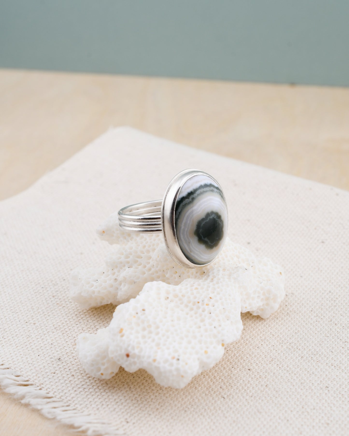 Silver ring with a green and white ocean jasper on a piece of white coral against a beige background