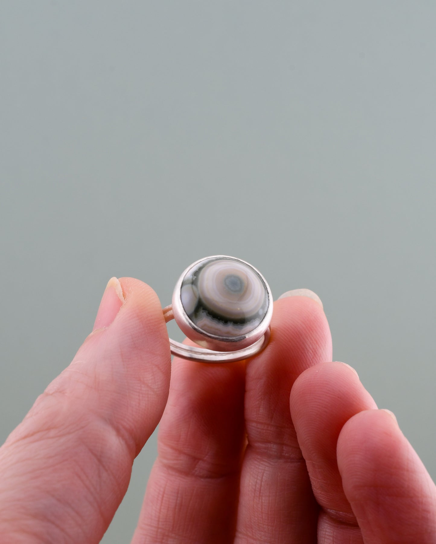 Silver ring with a round green and white ocean jasper held between fingers against a gray background