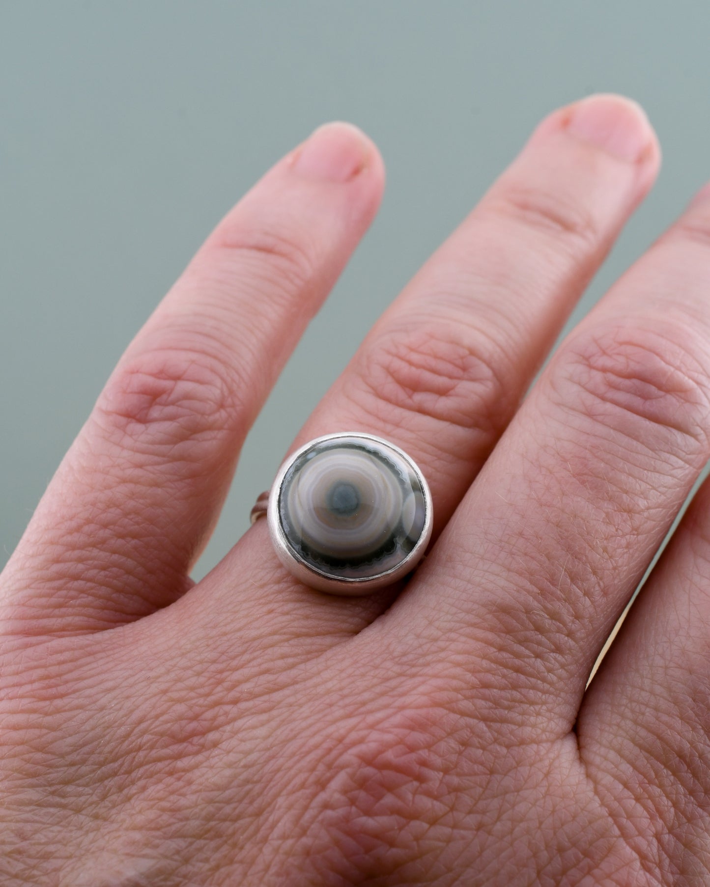 Hand wearing a silver ring with a jasper with white and green circular design on a gray background