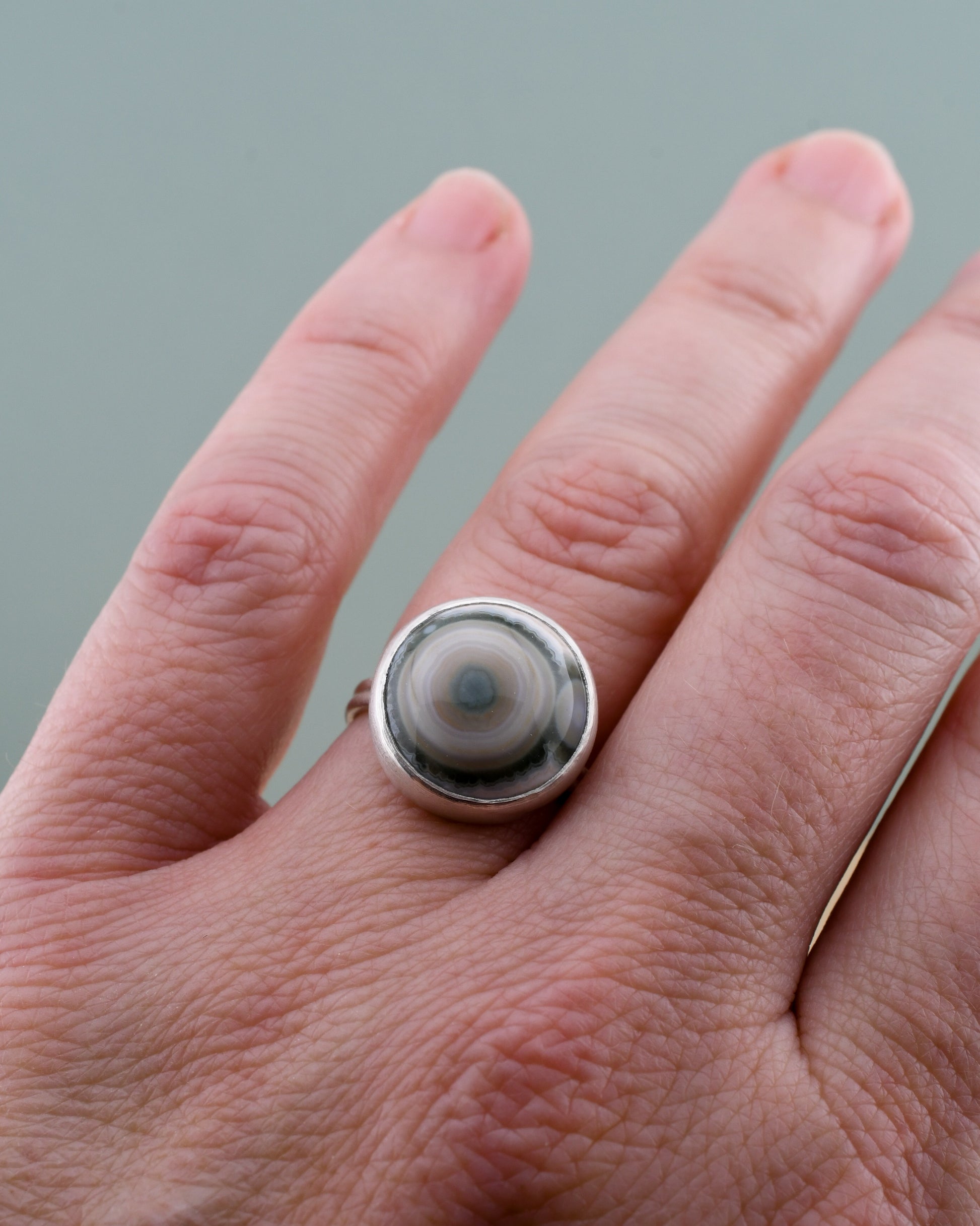 Hand wearing a silver ring with a jasper with white and green circular design on a gray background