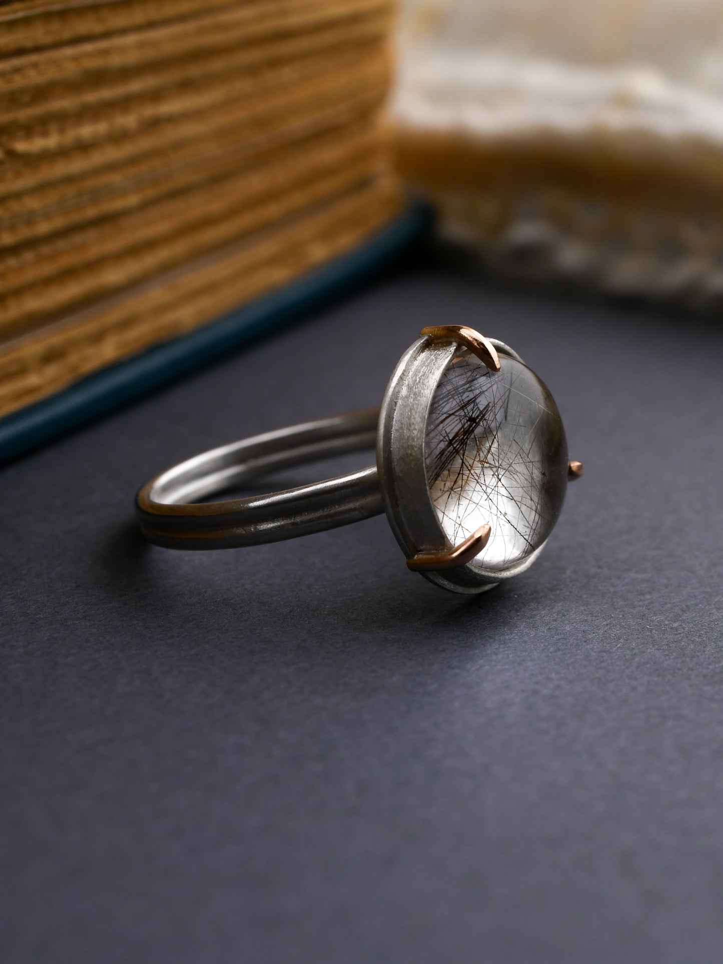 Silver ring with a central stone on a dark surface, with a book and fabric in the background.