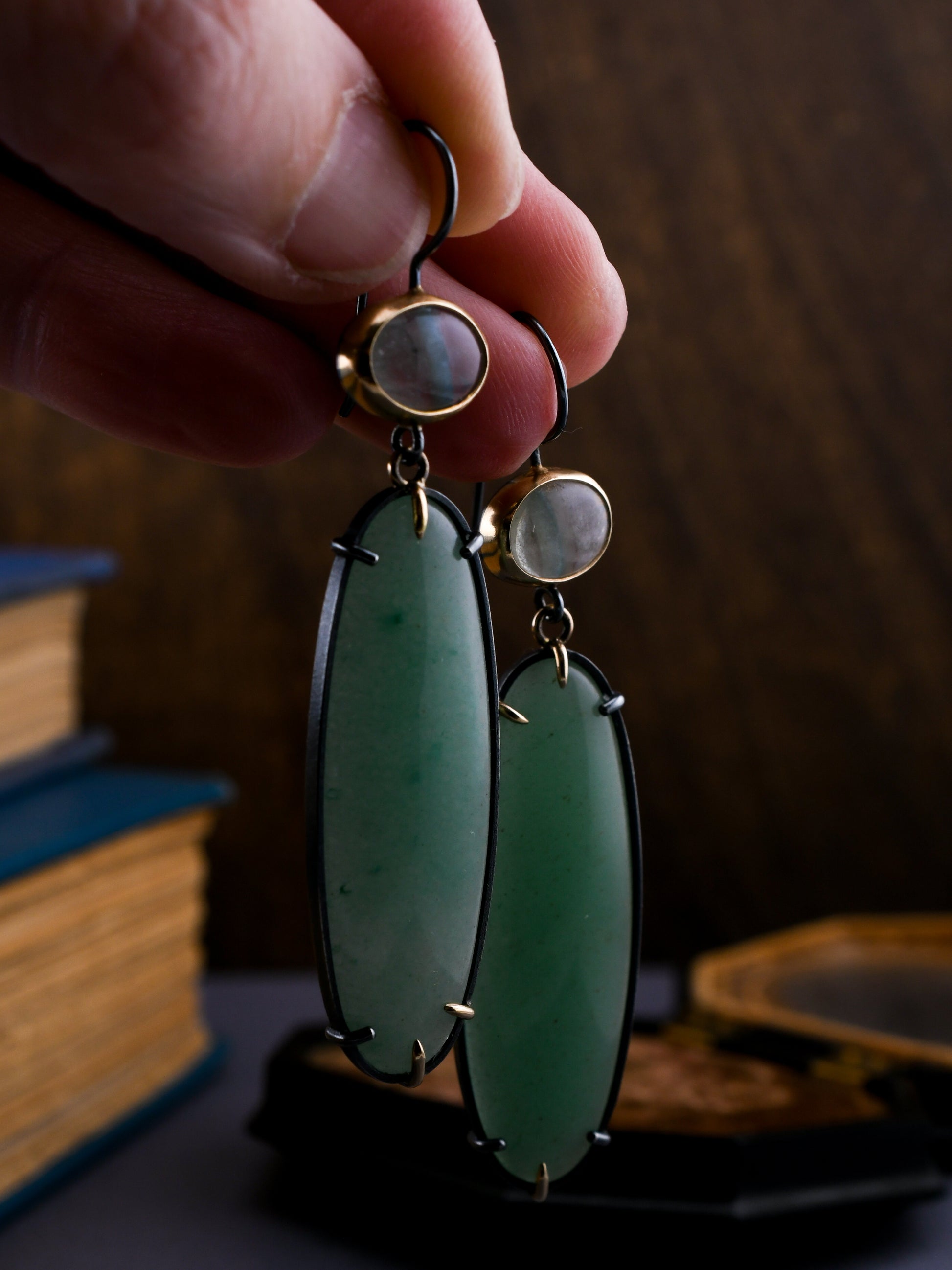 Hand holding a pair of earrings with green stones and gold accents against a blurred background.
