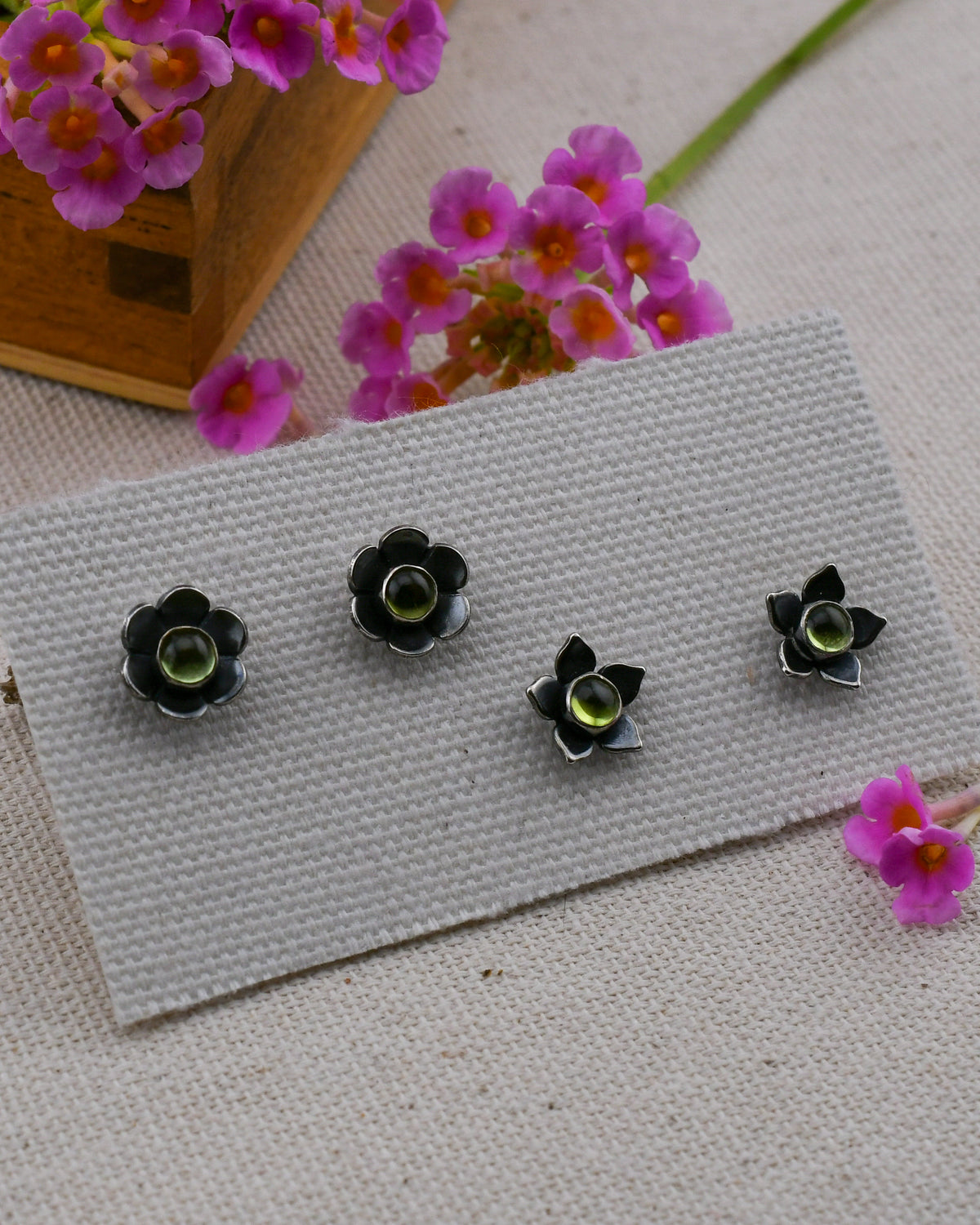 Two pairs of small black flower-shaped earrings with green peridot centers, arranged on a white textured card with pink flowers in the background.