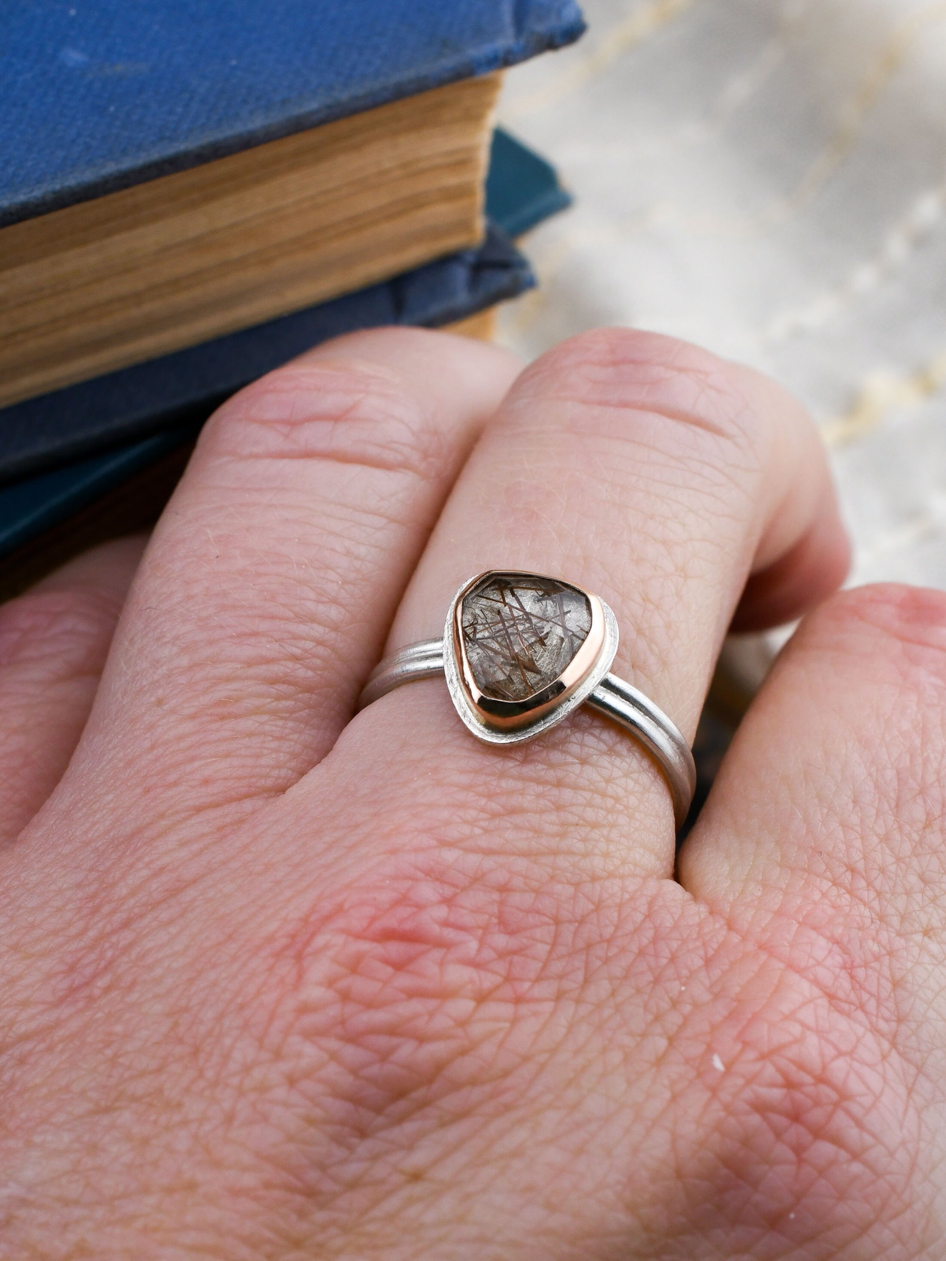 Hand wearing a silver ring with a heart-shaped gemstone against a book background