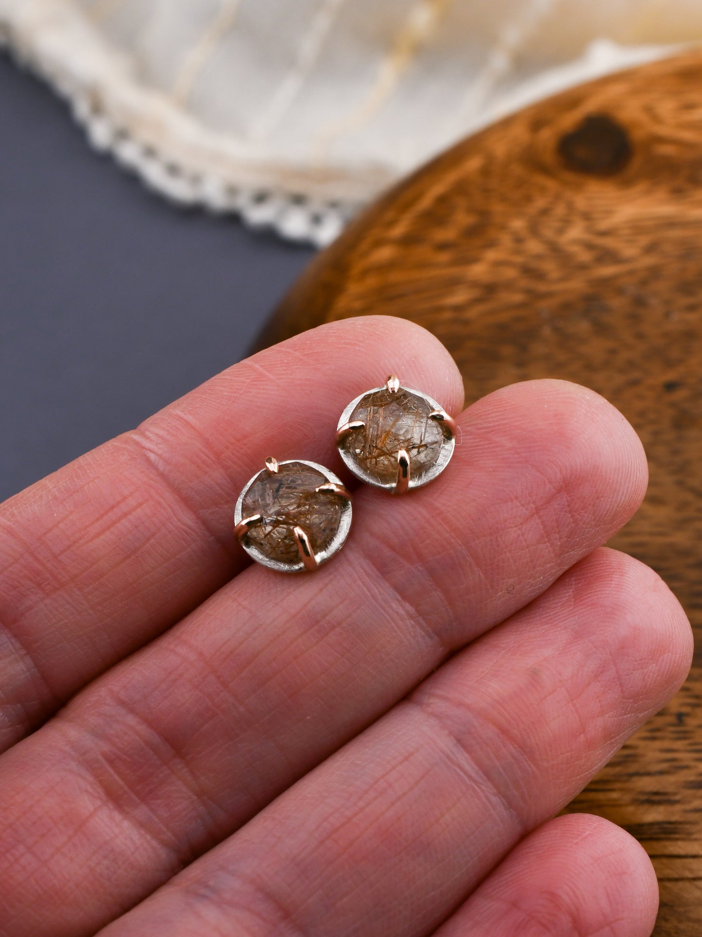 silver and rose gold rutilated quartz studs held in a hand on a dark background with wood and cream linens