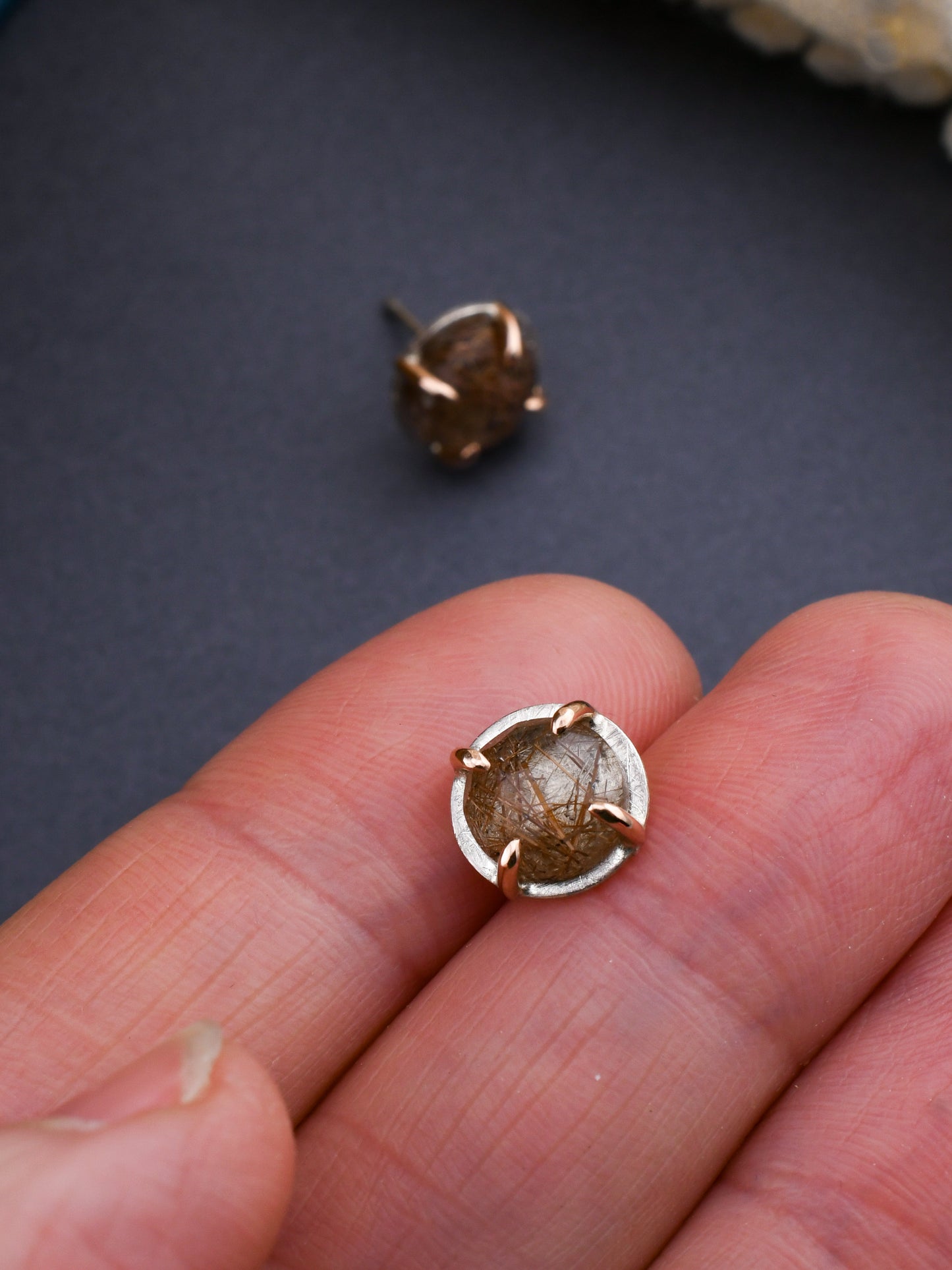 closeup of rutilated quartz stud earring held between fingers over a gray background