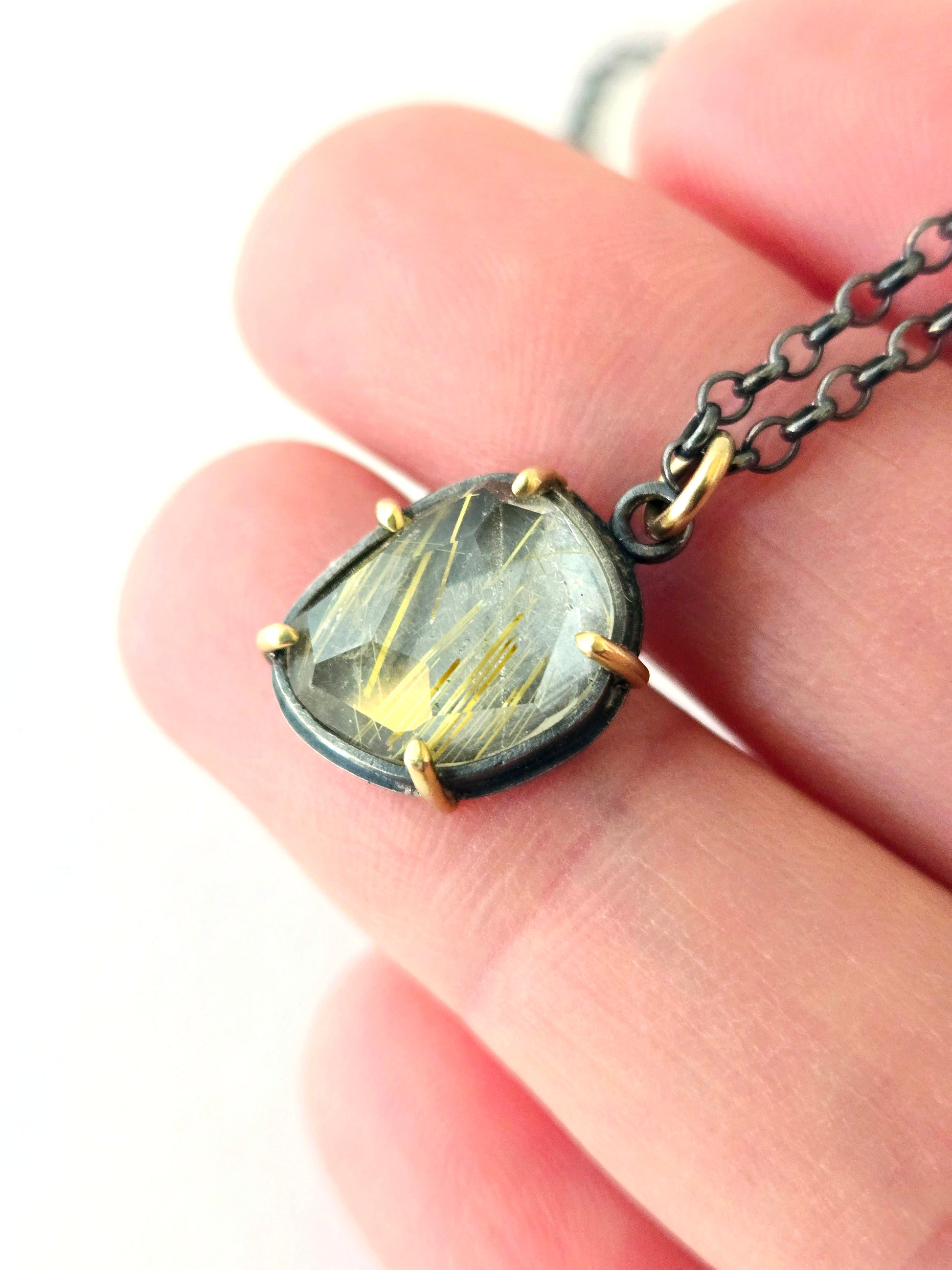 Closeup of rutilated quartz pendant set in 14k gold and blackened silver held in a hand over a white background