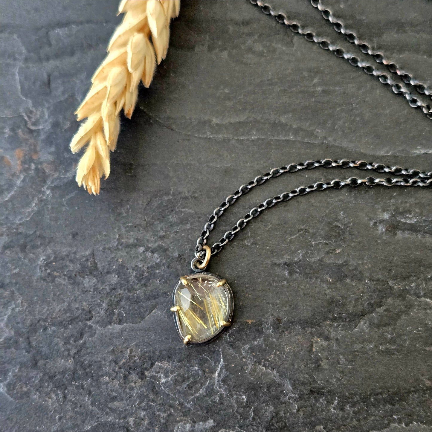 Black silver and yellow gold necklace with a golden rutilated quartz pendant on a textured stone surface with pale gold wheat