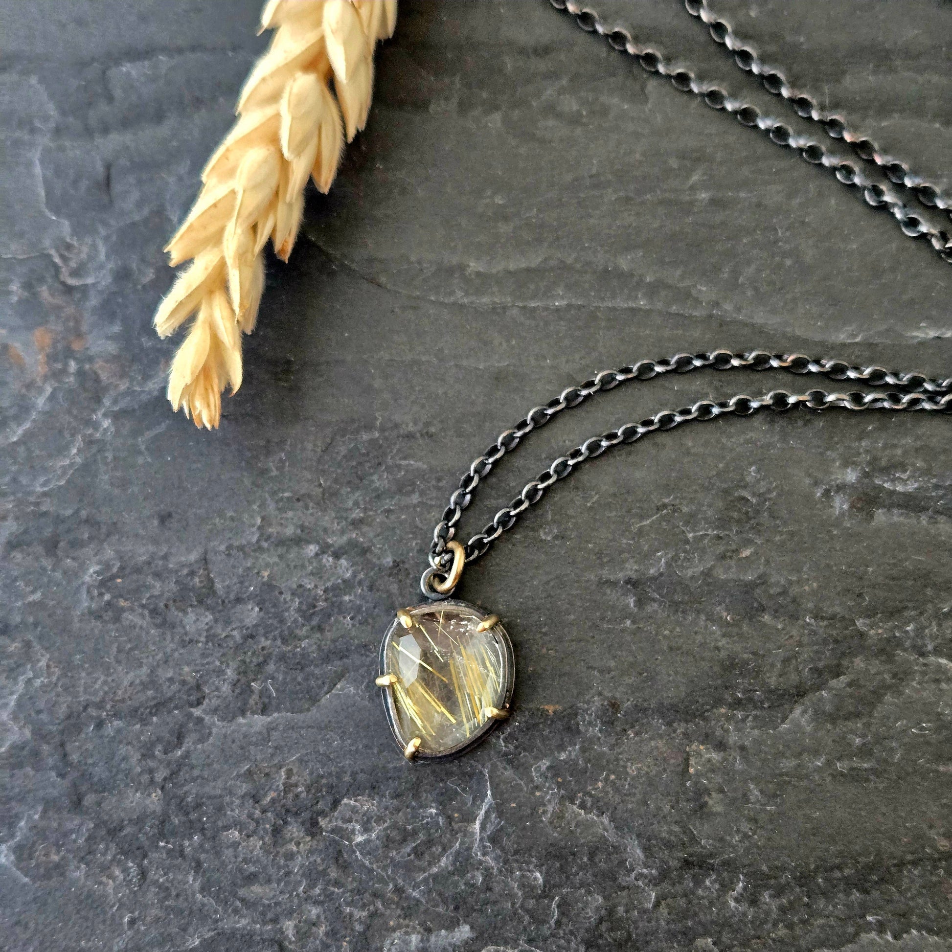 Black silver and yellow gold necklace with a golden rutilated quartz pendant on a textured stone surface with pale gold wheat