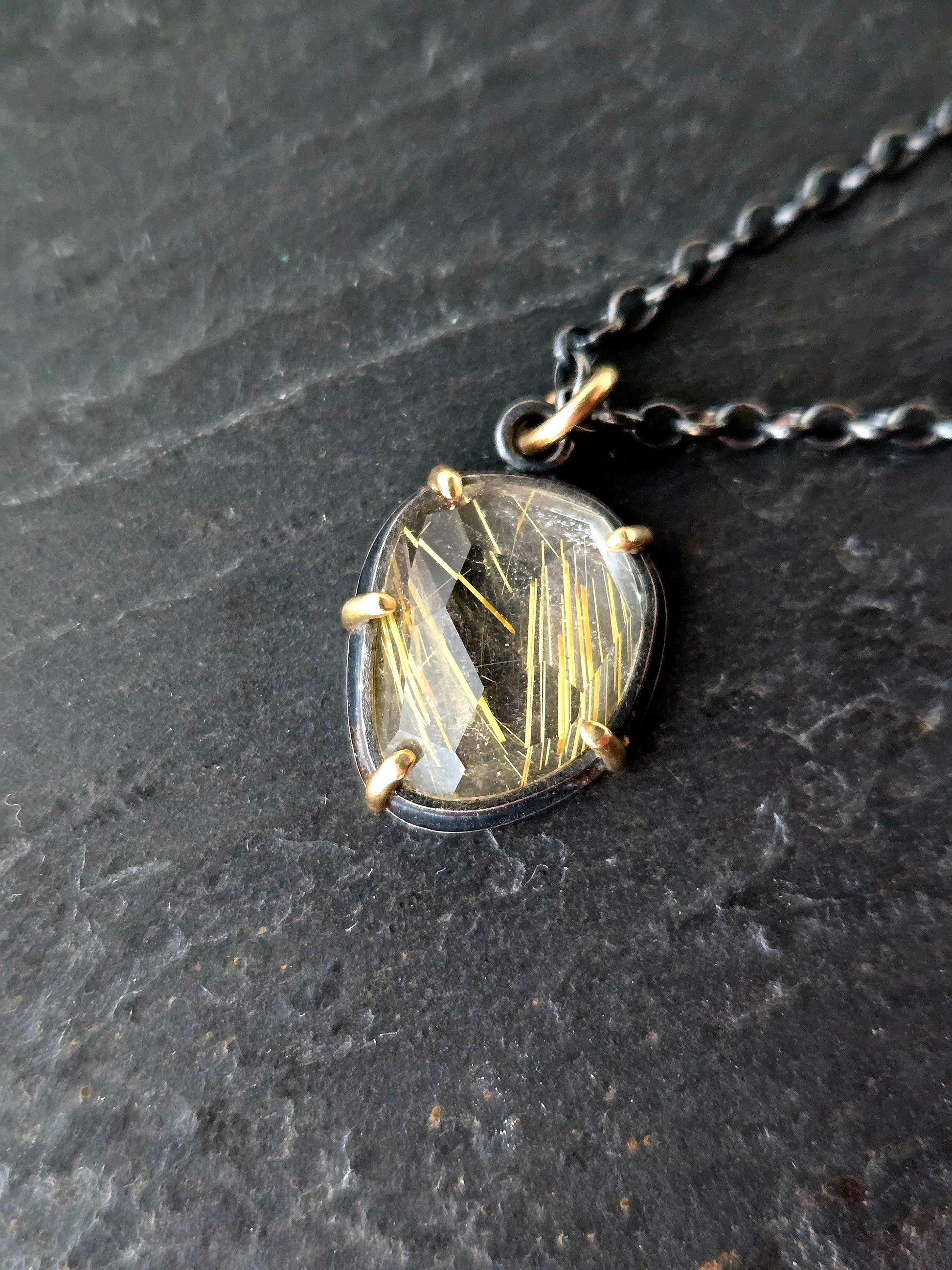 Closeup of golden rutilated quartz pendant in 14k gold and black silver on a textured black stone surface