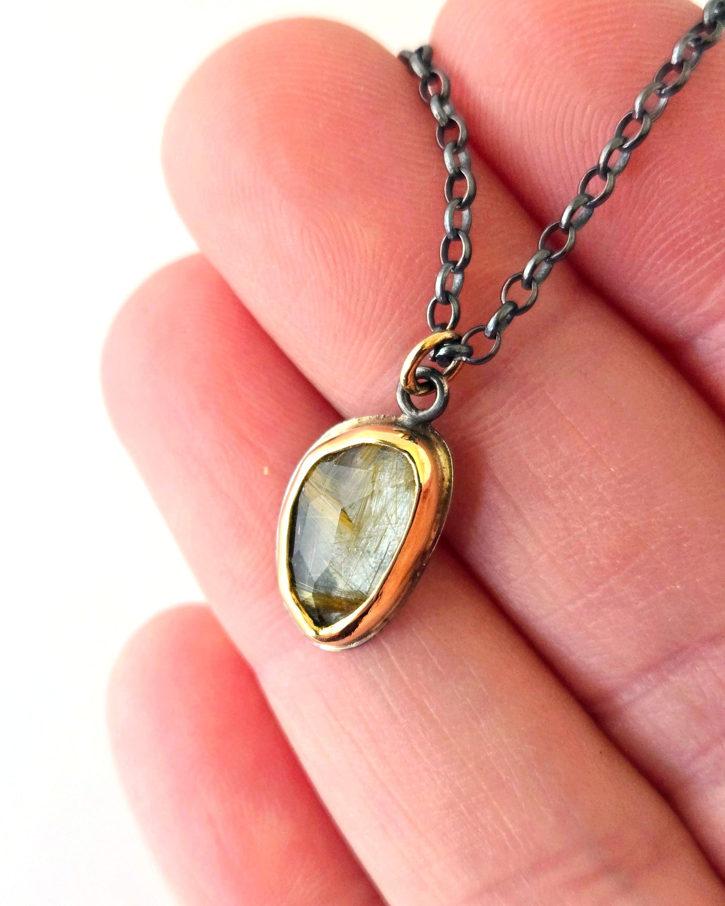 Necklace with a rutilated quartz pendant wrapped in yellow 14k gold held between fingers on a white background