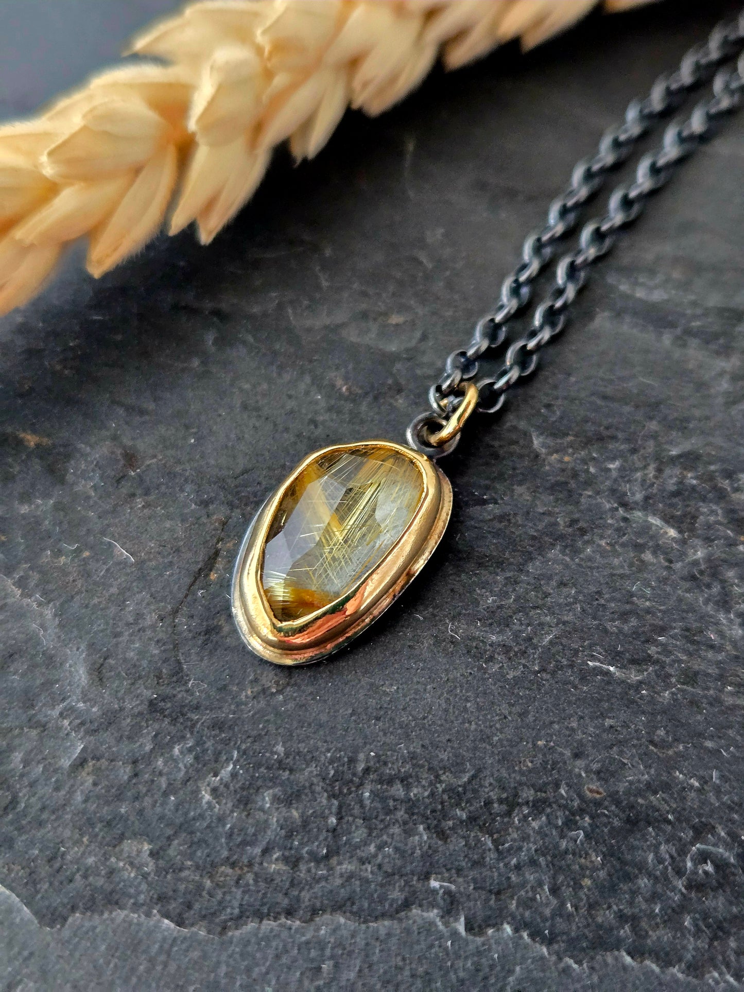 A pendant with a golden rose cut rutilated quartz set in a 14k gold bezel, attached to a silver chain, displayed against a stone background with wheat at the top.
