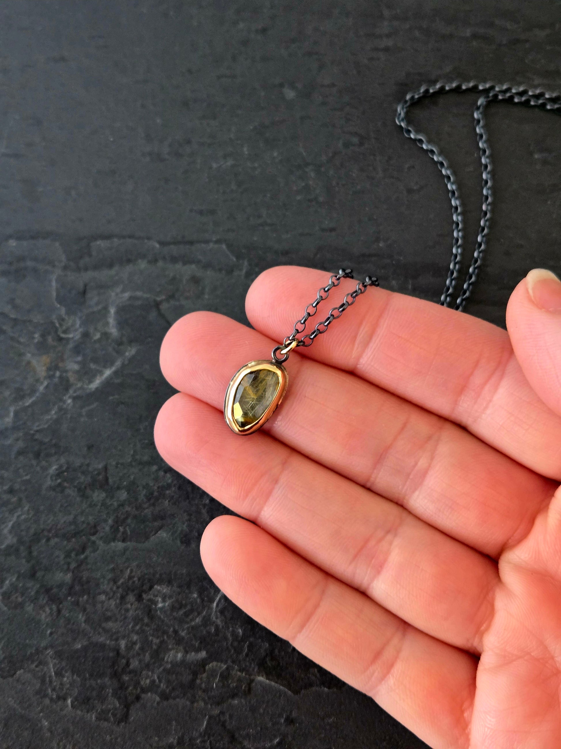 Rutilated quartz necklace in 14k gold and blackened silver held in a hand over a dark textured background