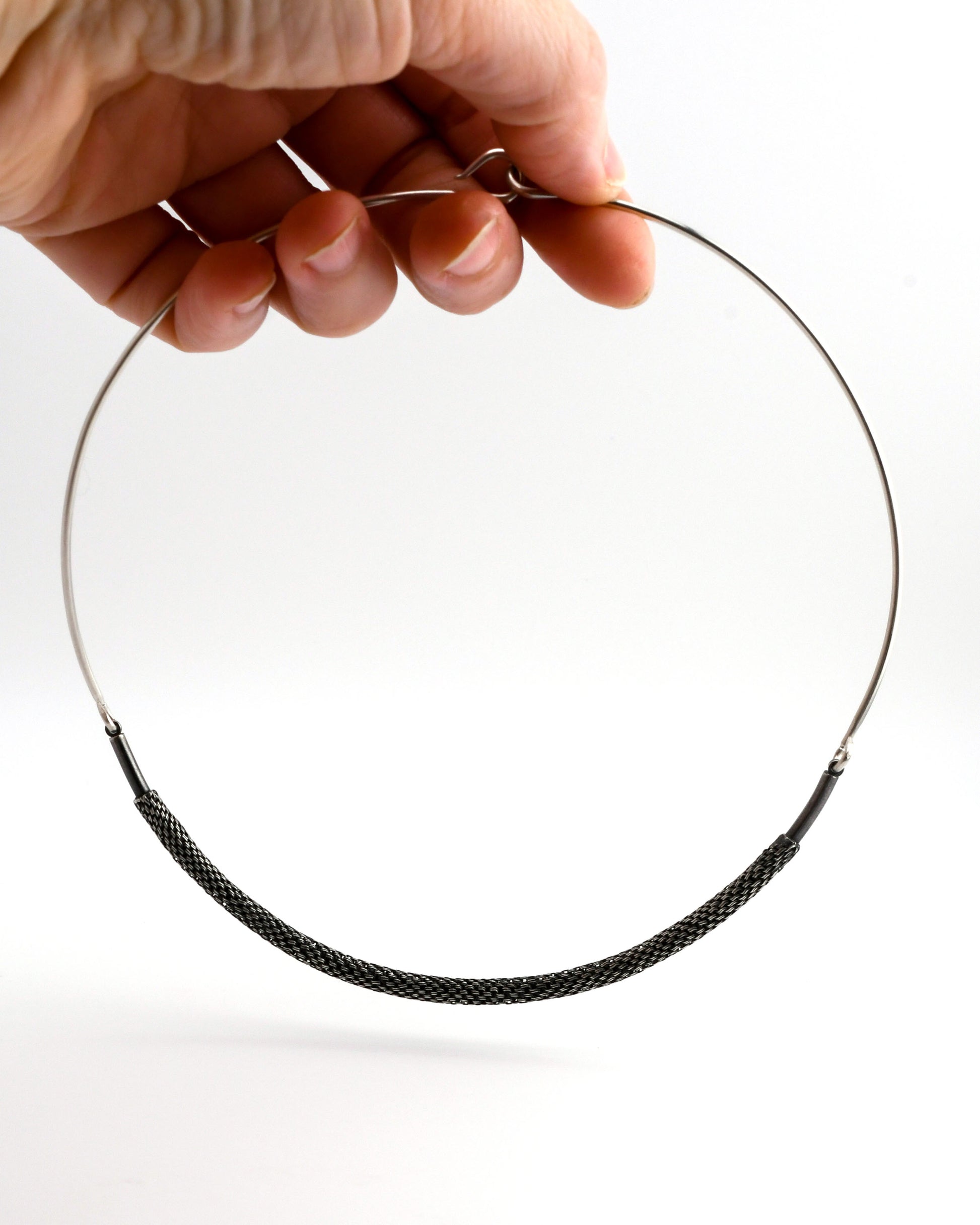 Woven silver torque necklace suspended from a hand on a white background