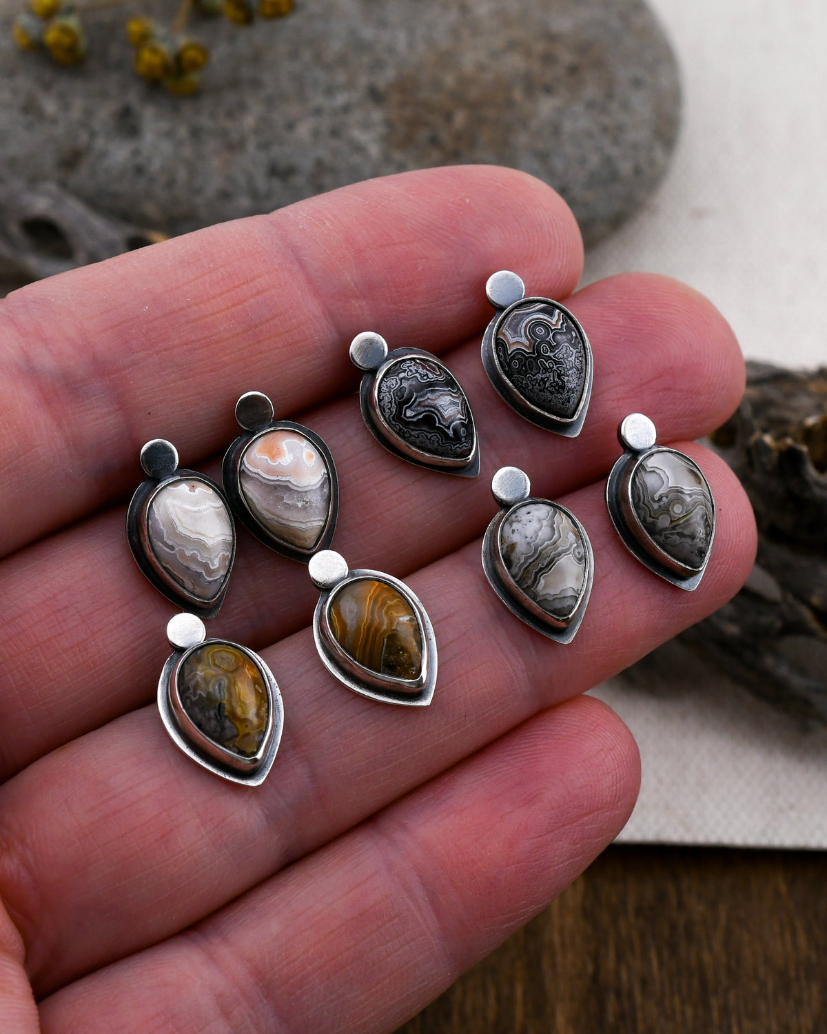 Four pairs of teardrop shaped stud earrings featuring crazy lace agate in different colors held between fingers on a natural background